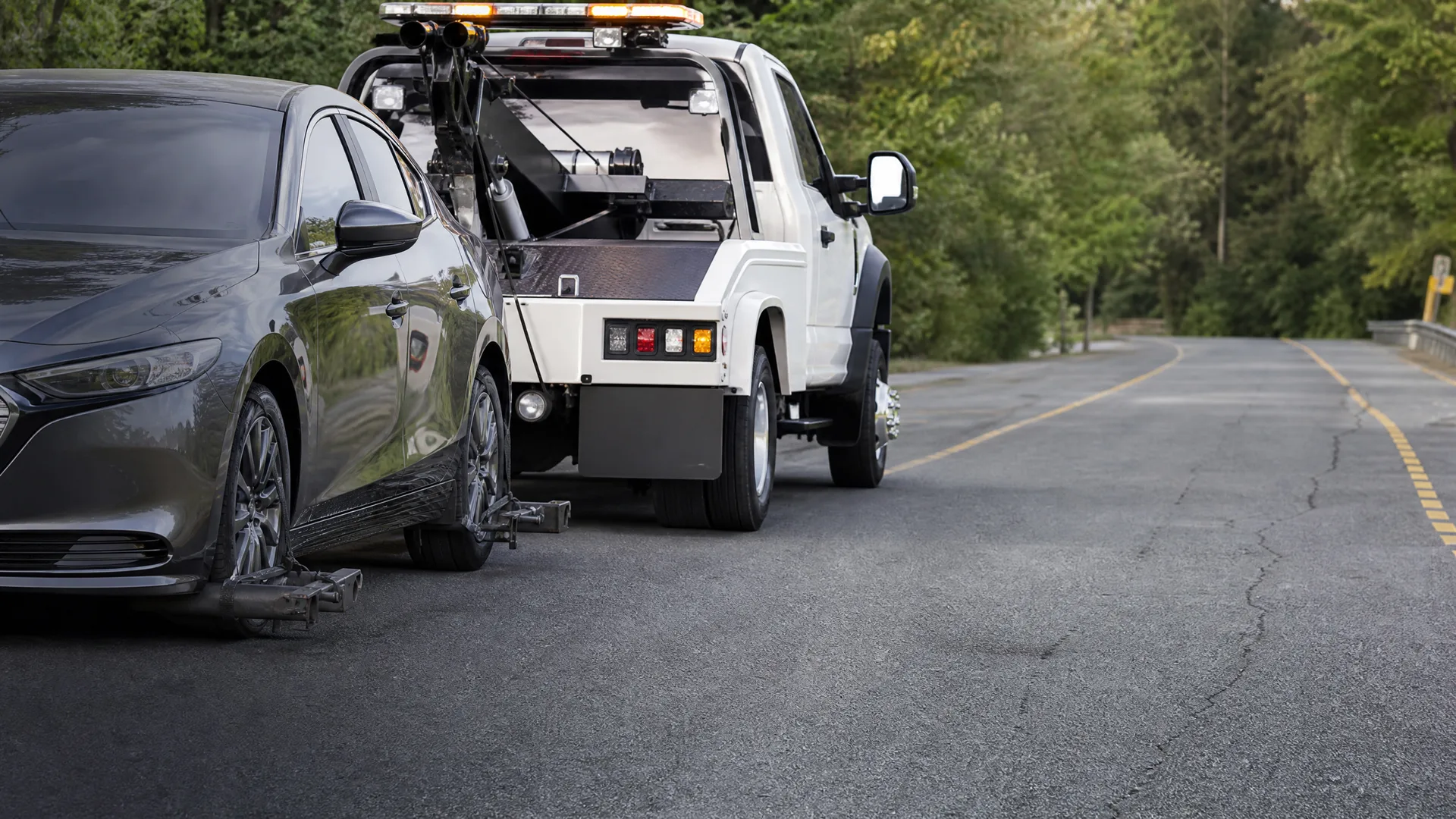 A white tow truck lifts the front wheels of a dark gray sedan on a paved road with a yellow center line, with green trees along the roadside.