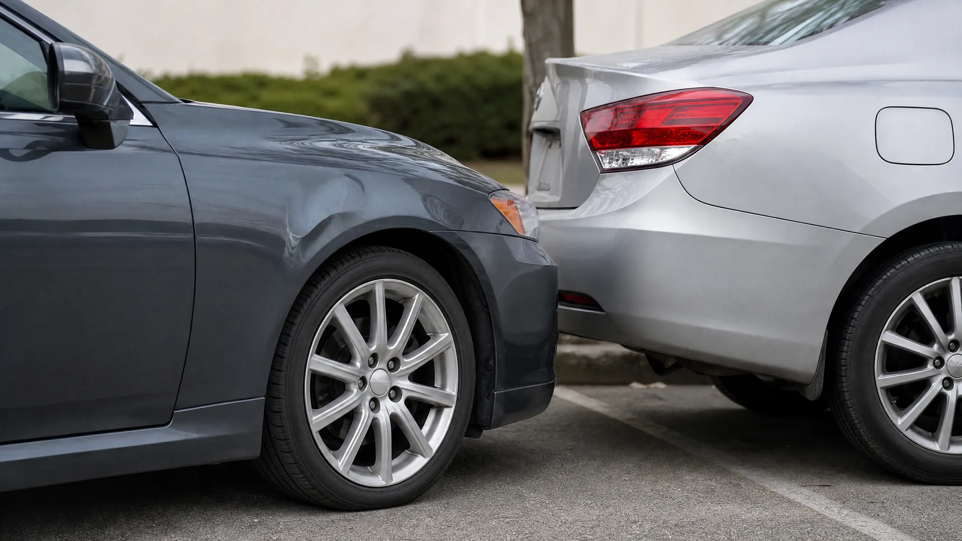 Close-up of the front corner of a dark gray sedan touching the rear bumper of a silver sedan in a paved parking area with hedges and a wall behind.