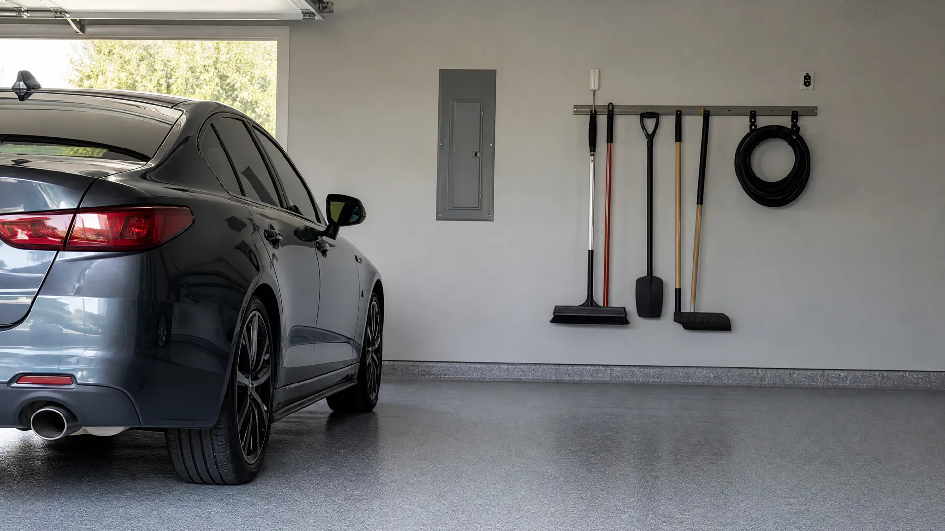 A dark sedan parked in a garage with a speckled epoxy floor, beside a gray wall holding an electrical panel and a rail with hanging brooms, a shovel, and a coiled hose.