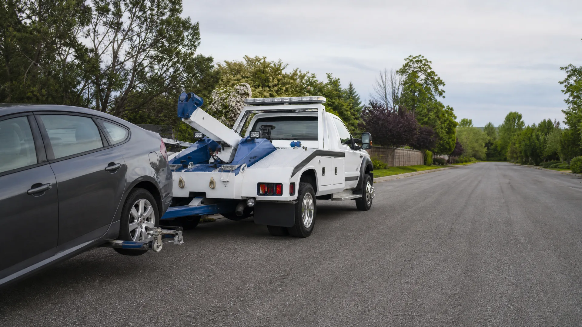 A white and blue tow truck towing a gray sedan by its front wheels on a paved residential street lined with trees.
