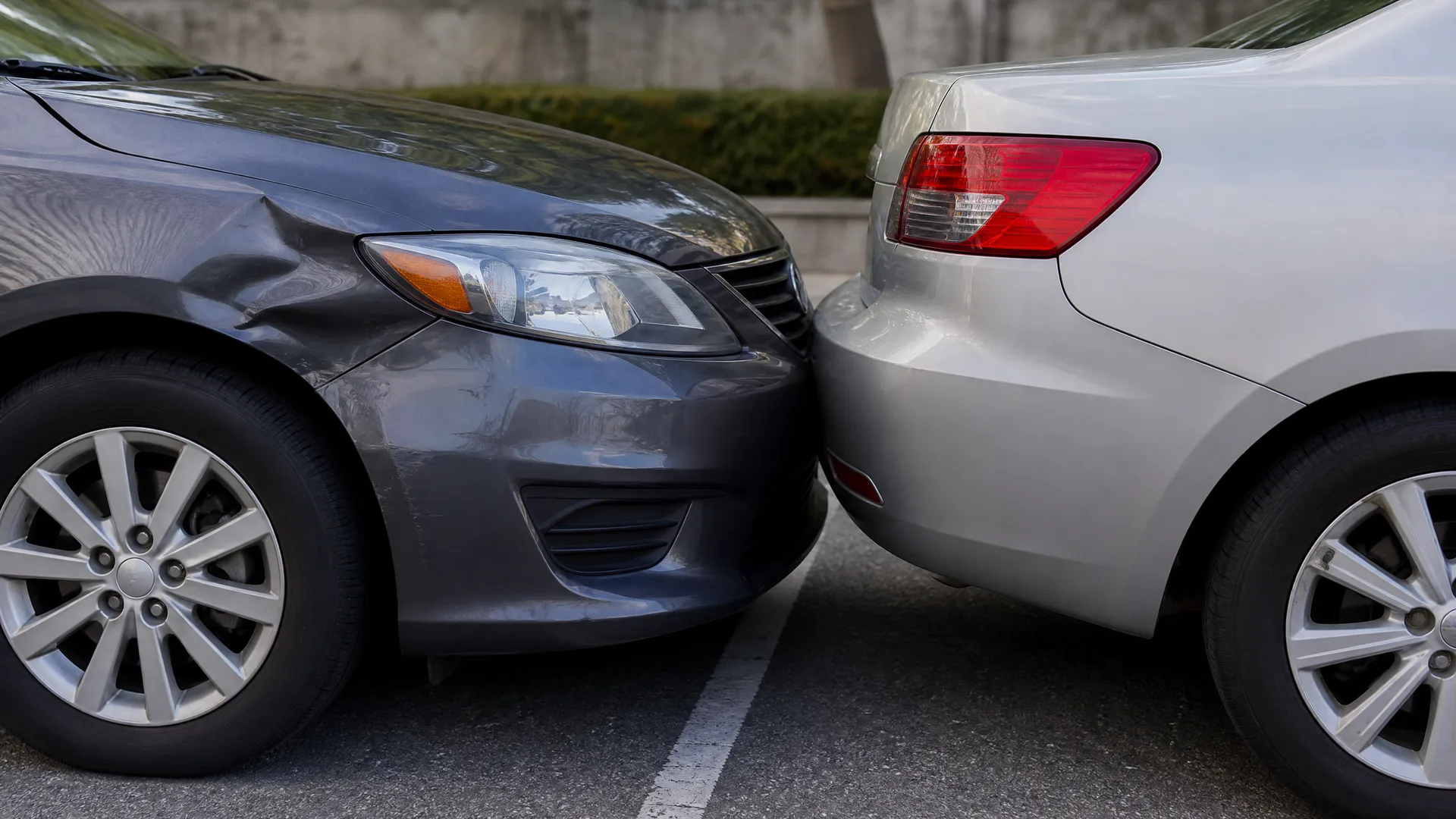 Front bumper of a dark gray sedan with a dented fender touching the rear bumper of a silver sedan in a parking lot, with a hedge and wall in the background.
