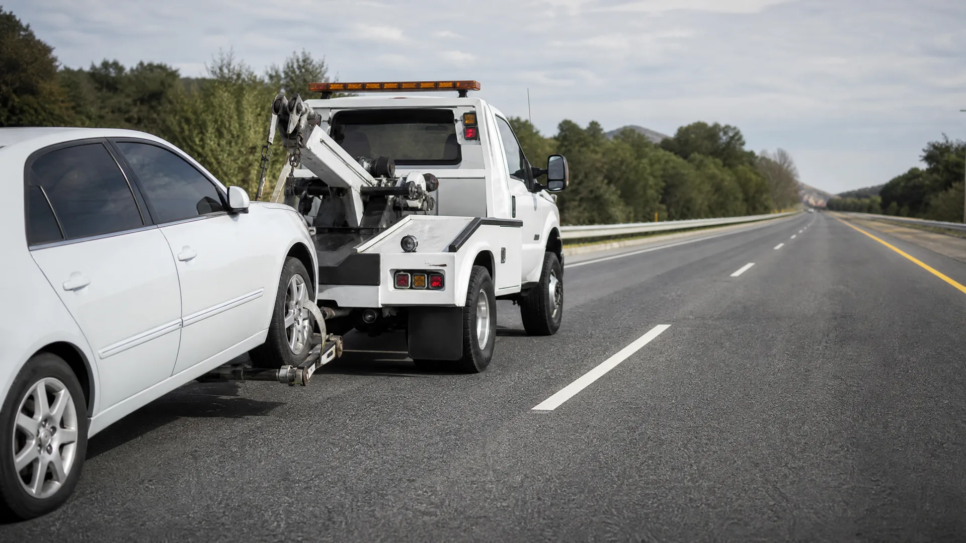 A white tow truck on the shoulder of a two-lane highway towing a white sedan by its front wheels, with trees and distant hills in the background.