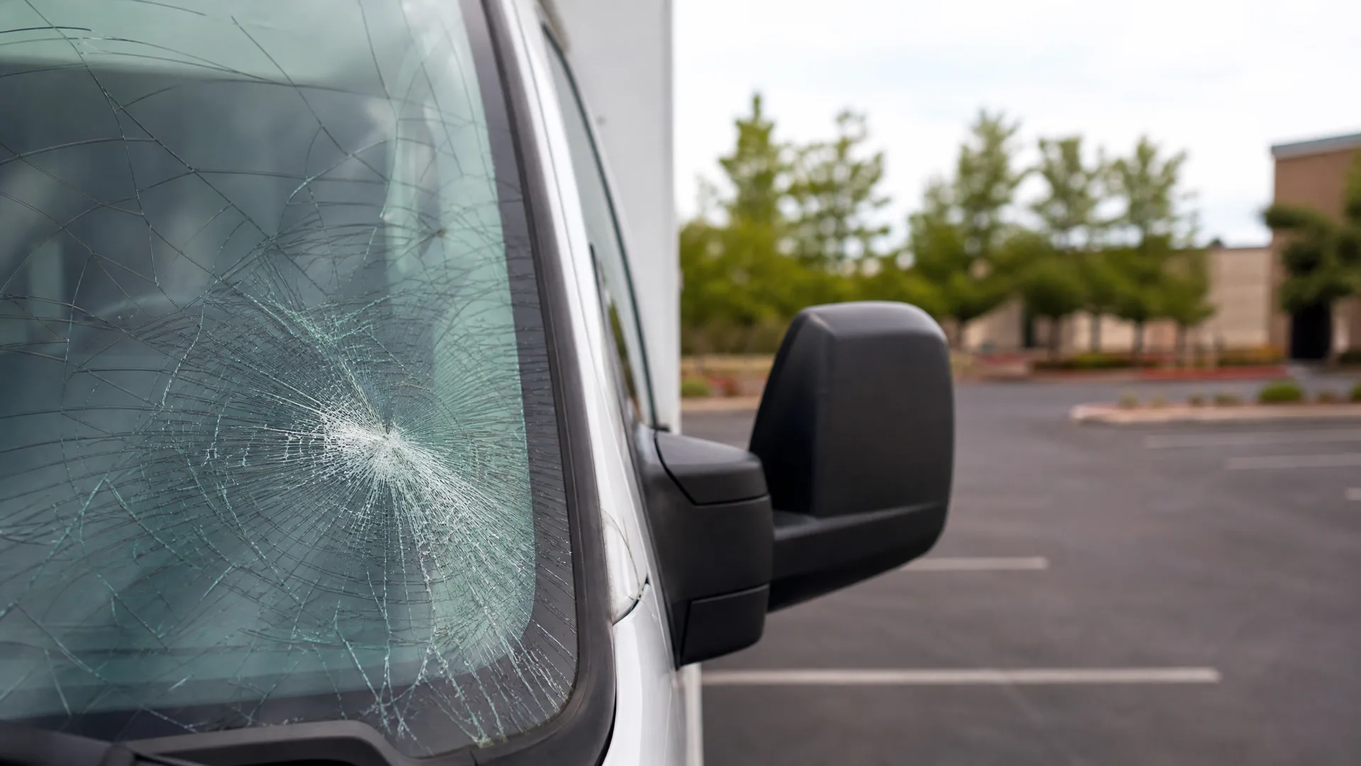Close-up of a white van's heavily cracked windshield with a central impact point, with the black side mirror and an empty parking lot with trees and a building in the background.