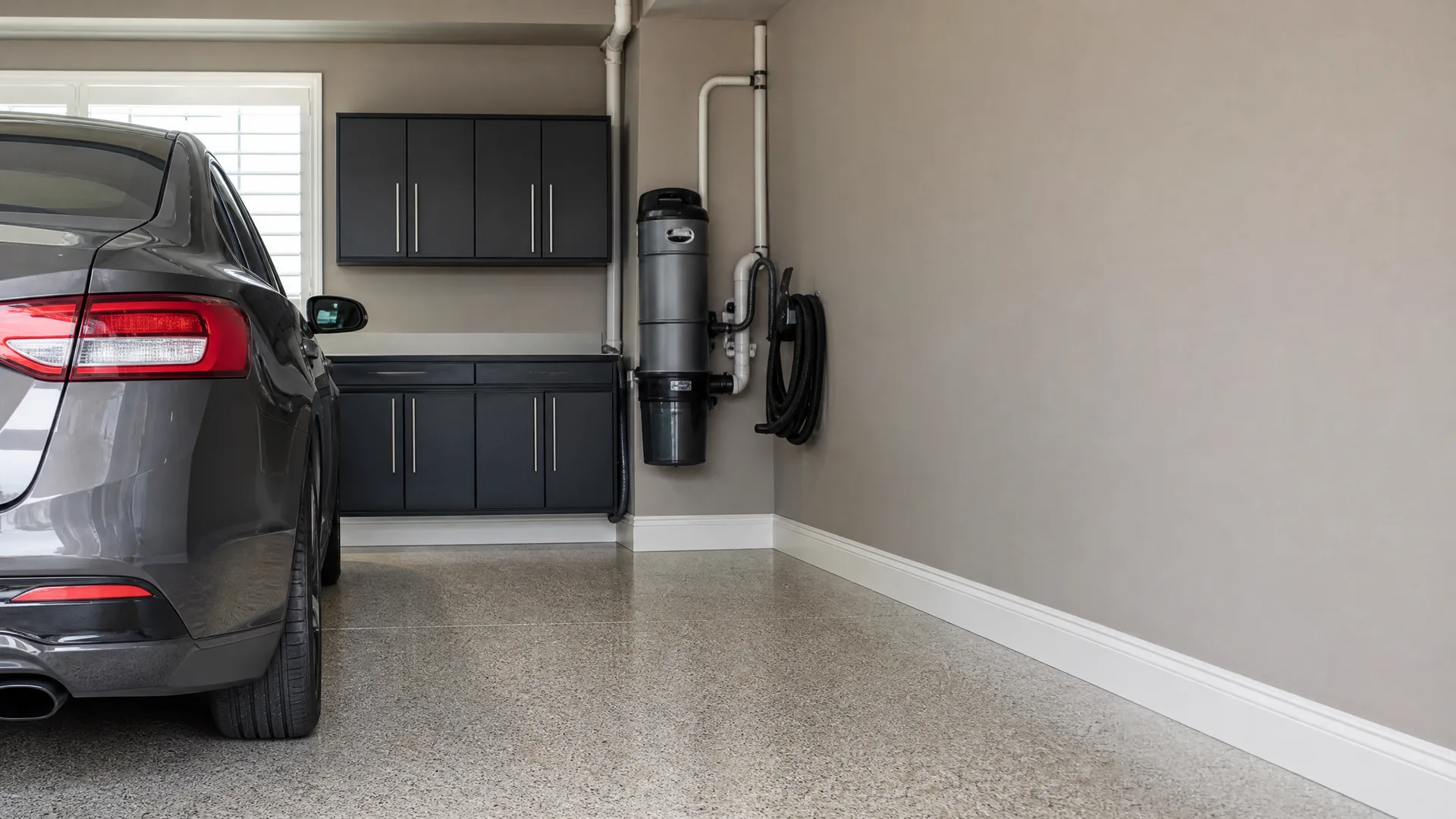 Garage interior with a polished speckled concrete floor, a dark gray sedan parked at left beside black cabinets, and a wall-mounted central vacuum canister with piping and a coiled hose on the beige wall.