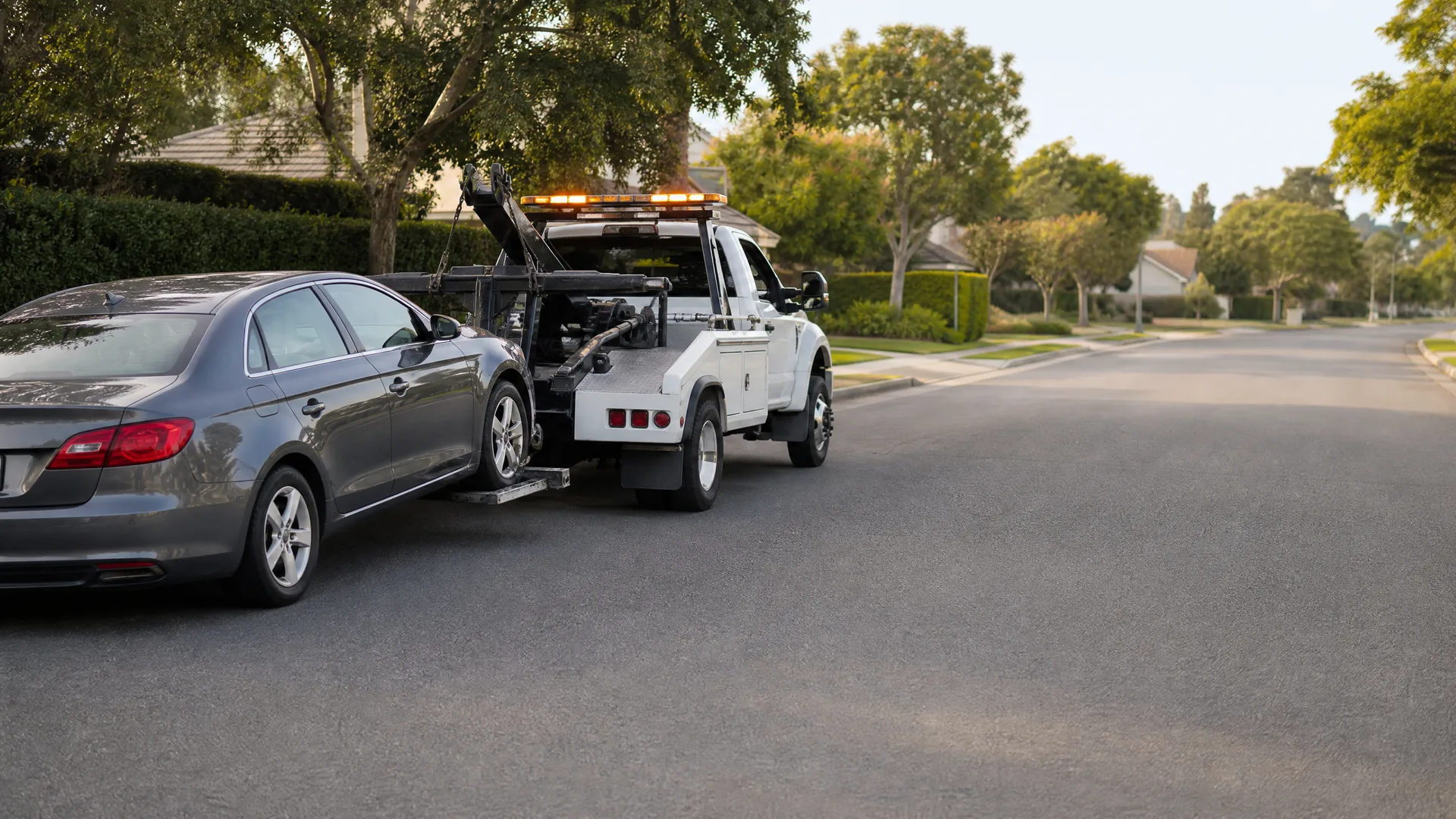 A white tow truck lifts the front of a gray sedan on a residential street lined with trees and hedges.