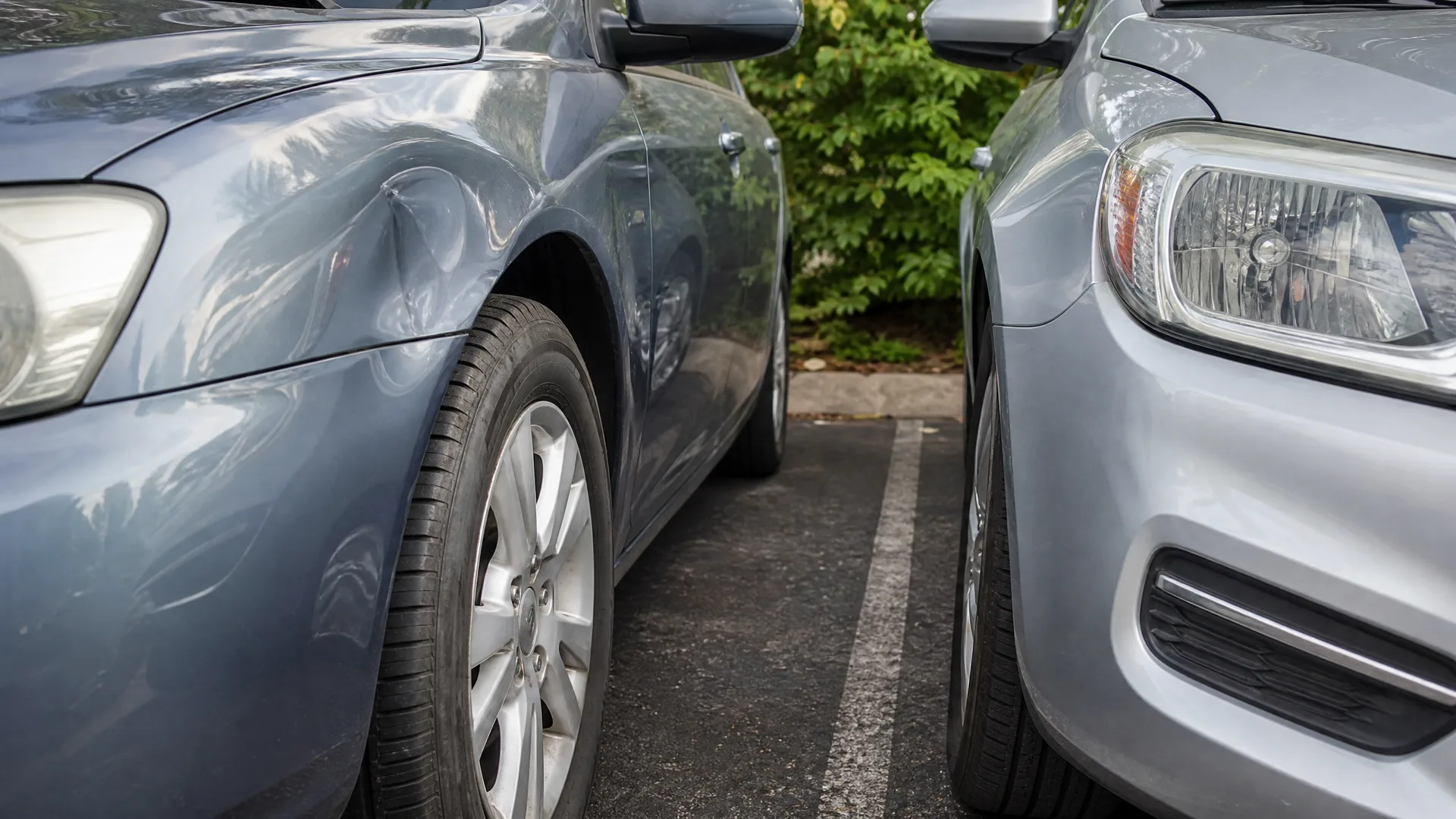Two cars parked closely side by side on asphalt with a painted white line between them, the dark blue car on the left showing a dent in its front fender, with green shrubs behind.