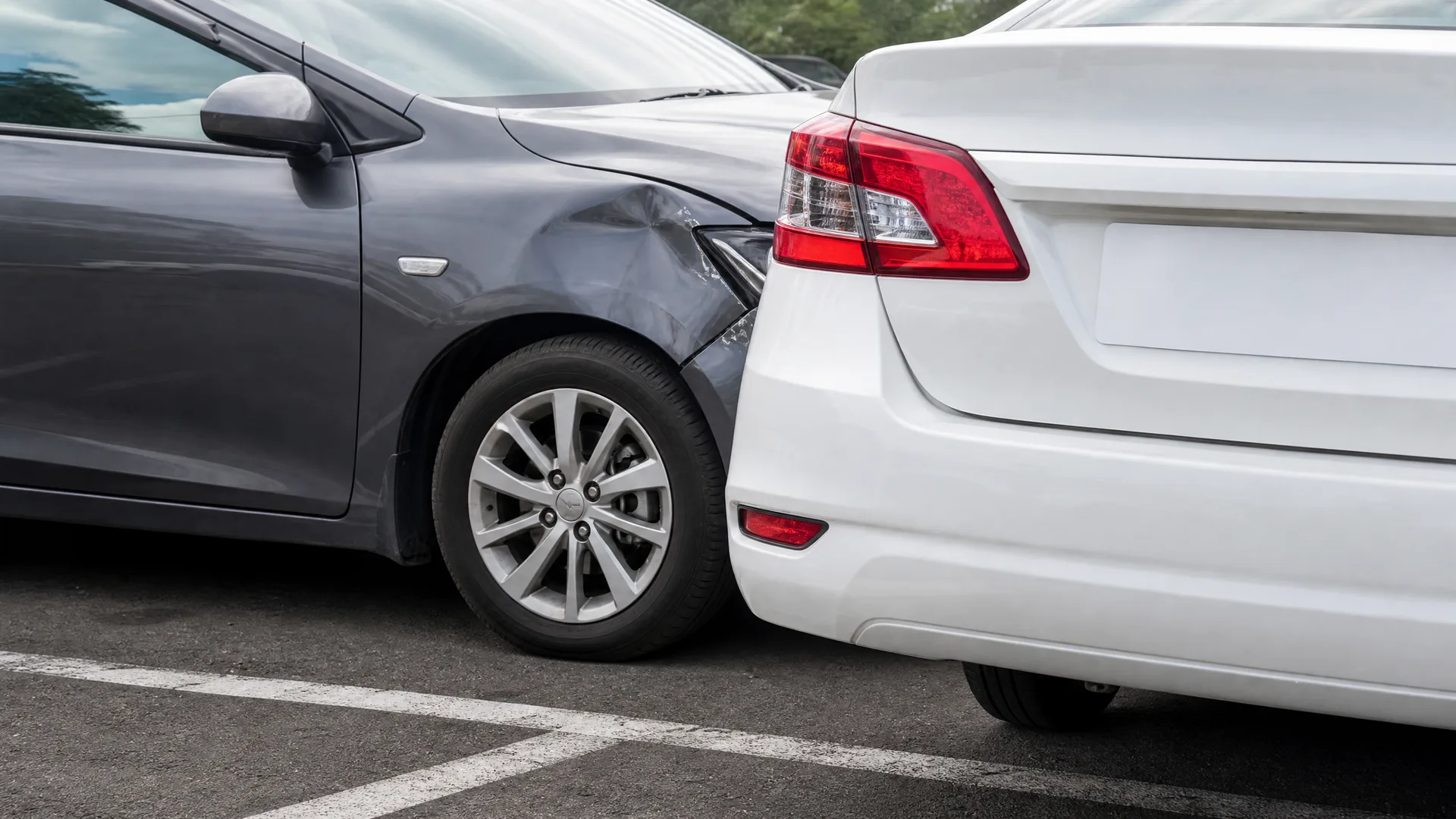A gray car's dented front fender and wheel pressed against the rear bumper and taillight of a white sedan in a paved parking lot with white line markings.