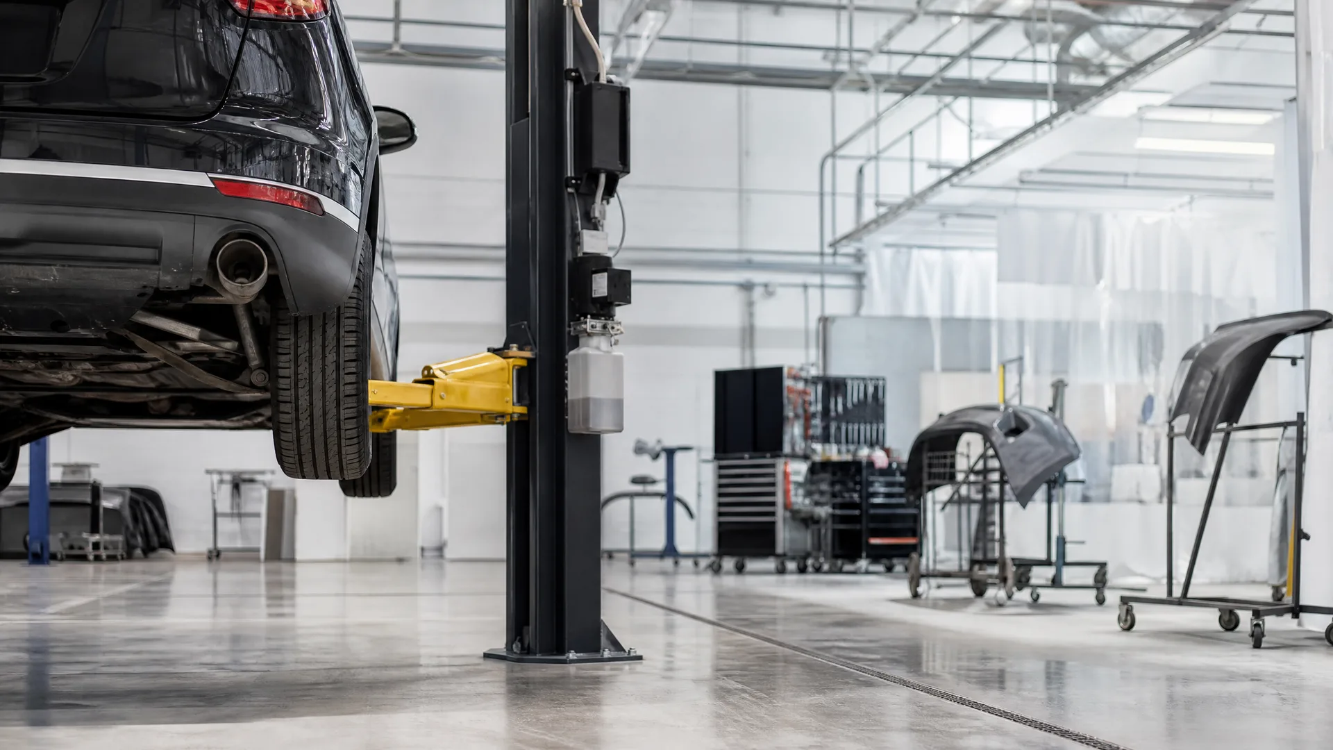 A dark car raised on a two-post lift with yellow arms inside a bright workshop, with tool cabinets, body panels on rolling racks, and a polished concrete floor in the background.