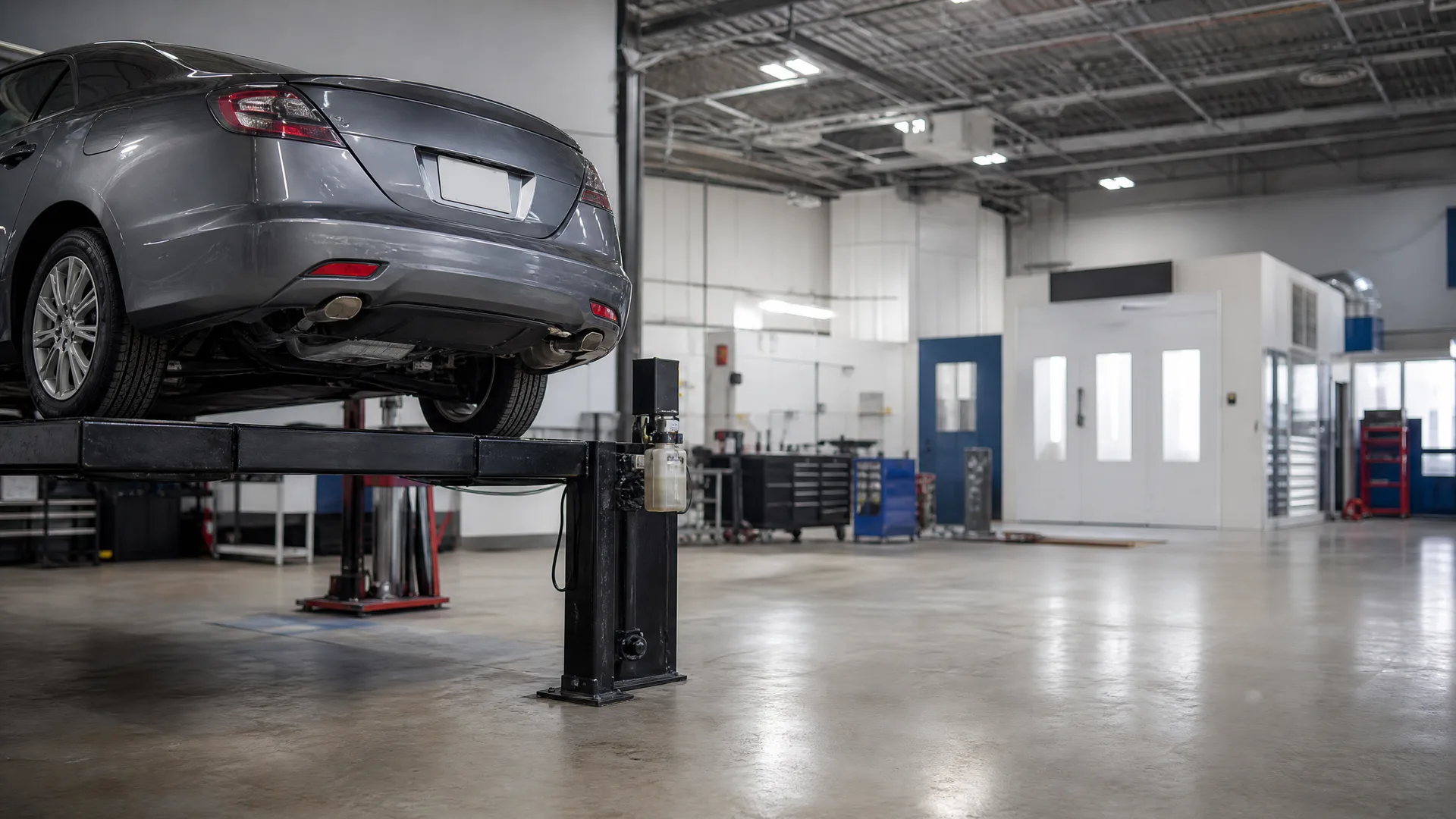 A gray sedan raised on a two-post vehicle lift inside an auto repair shop with a polished concrete floor, tool cabinets, and a white spray booth in the background.