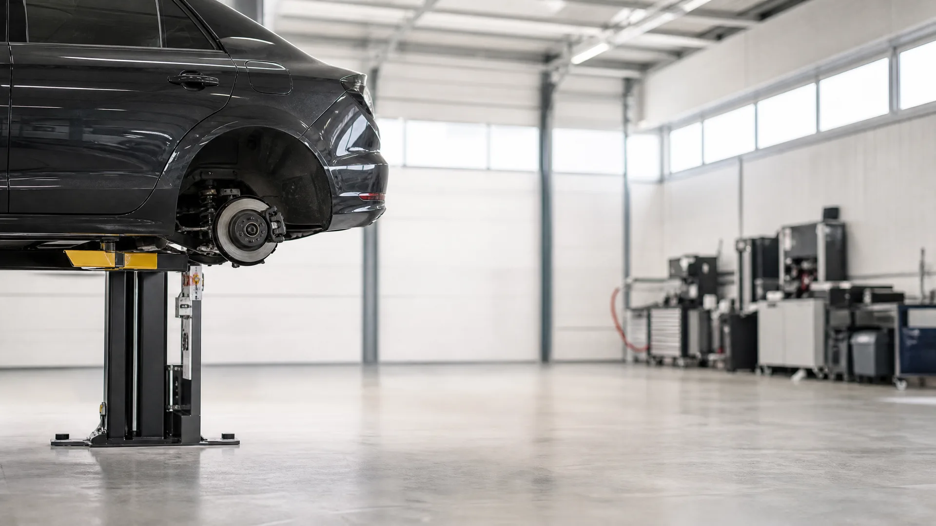 A dark car raised on a two-post lift with its rear wheel removed, inside a bright garage workshop with a polished concrete floor and tool cabinets along the far wall.