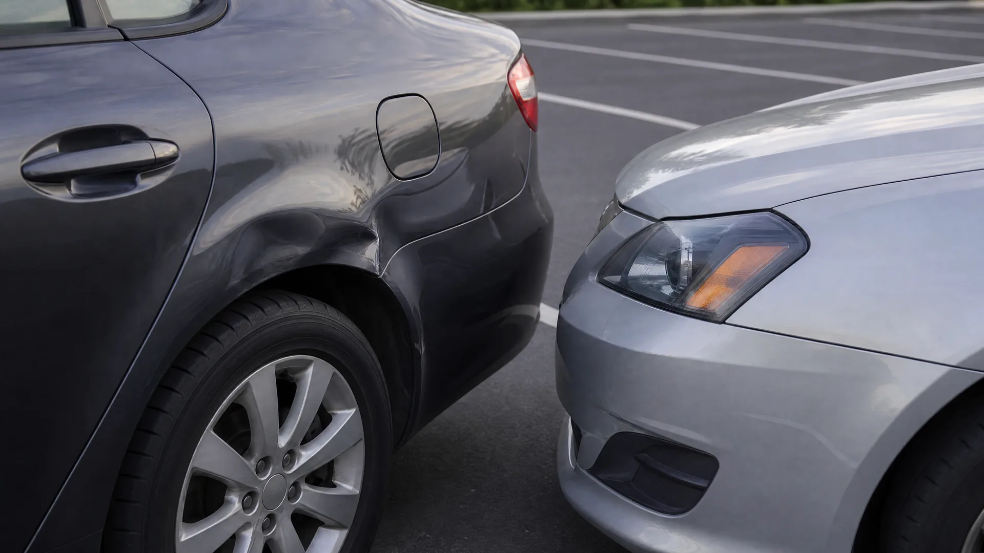 Close-up of a silver car's front end touching the dented rear side panel of a dark gray car in a parking lot with white painted lines.