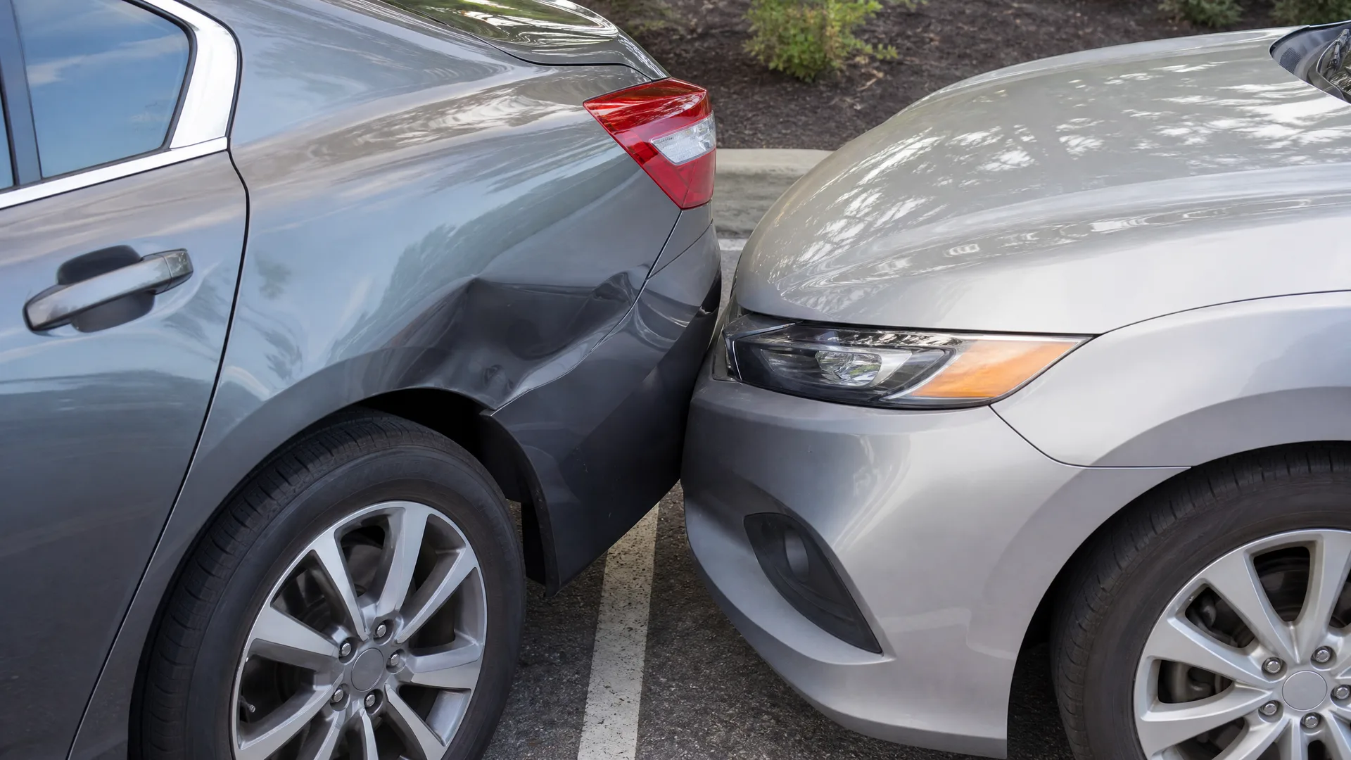 Close-up of two cars in a parking lot touching bumper to headlight, with a dent in the gray car's rear quarter panel.