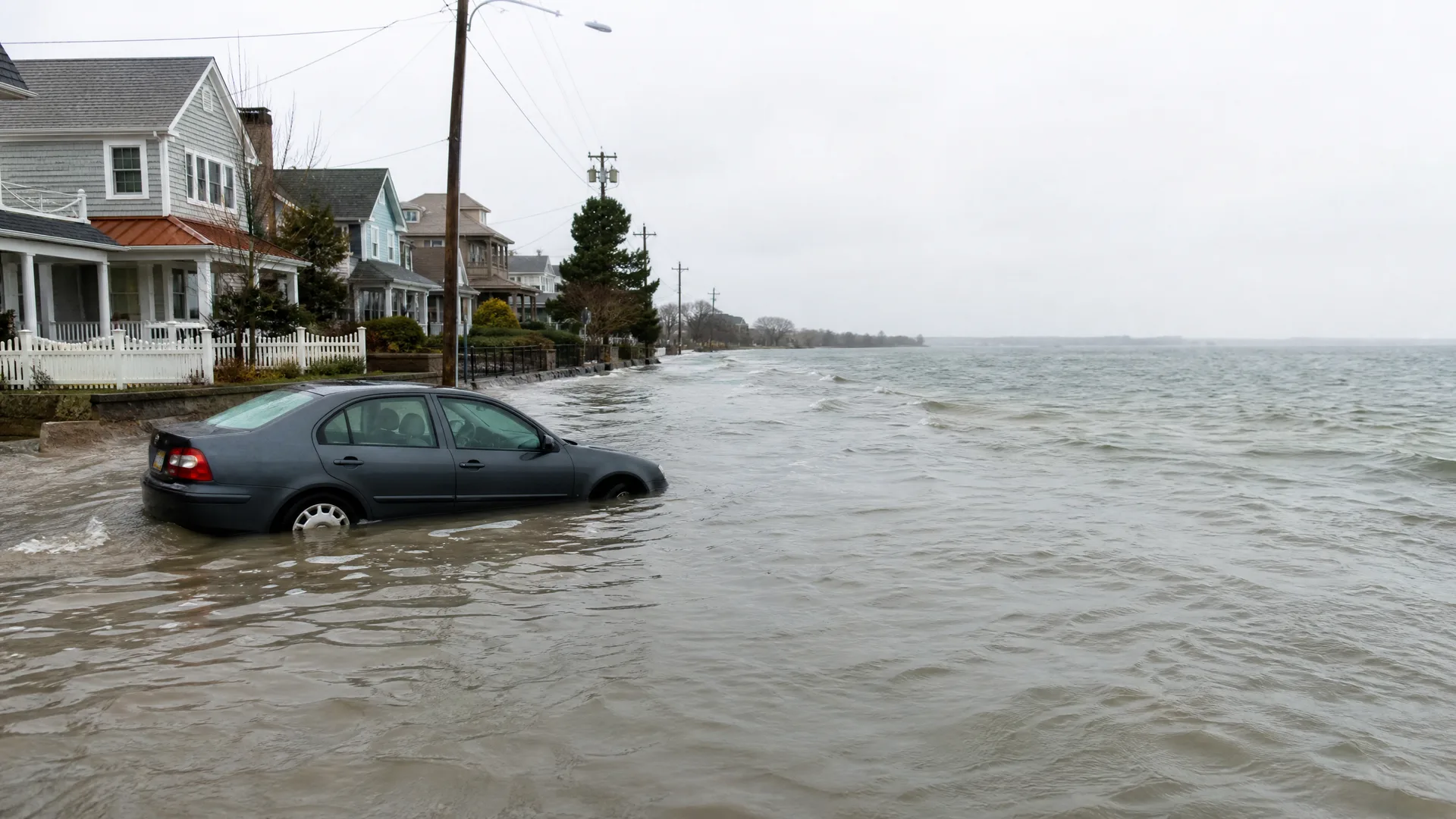 A dark sedan sits partly submerged in floodwater covering a coastal street beside white clapboard houses, with open water and a grey sky in the background.
