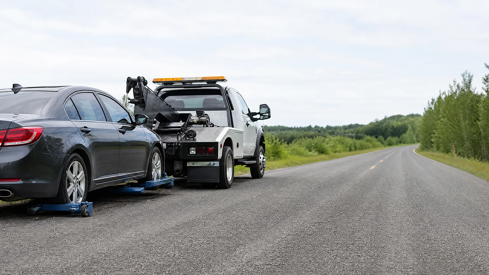 A white tow truck with an amber light bar towing a dark gray sedan on wheel dollies at the edge of a rural two-lane road lined with green trees.
