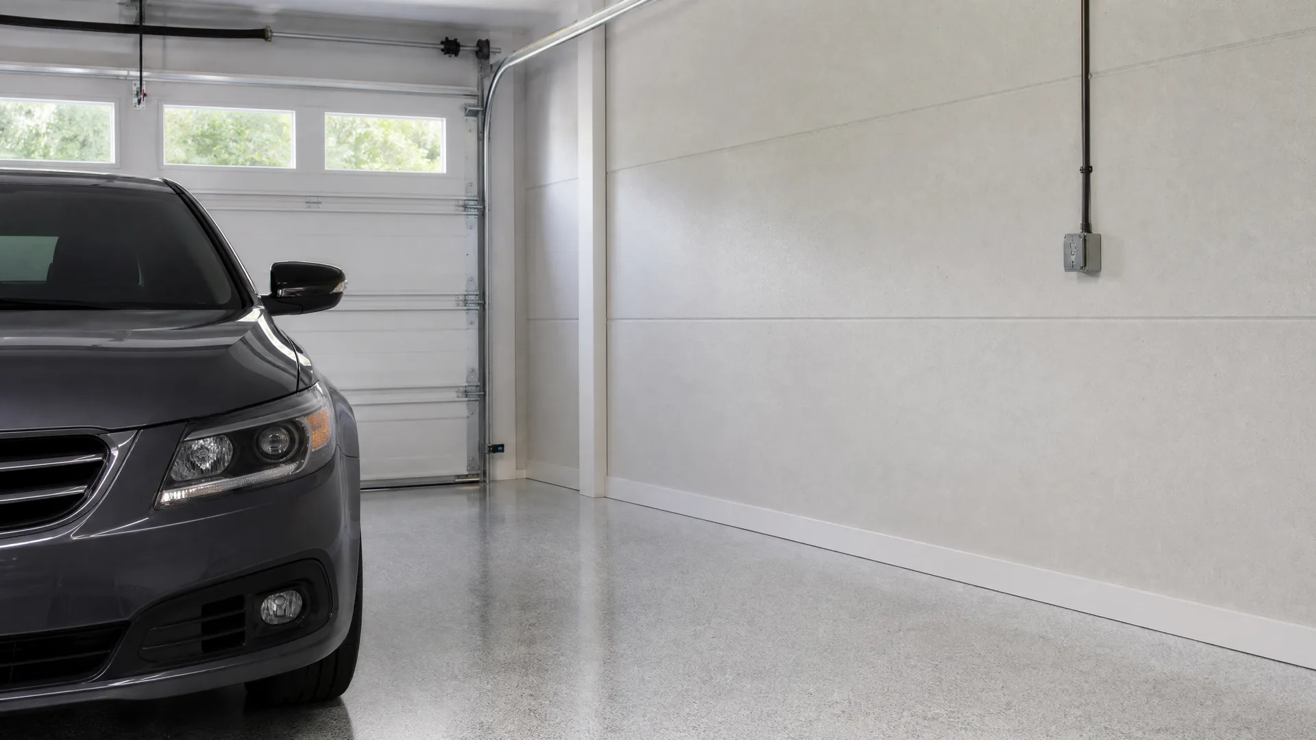 Interior of a garage with a polished concrete floor, a white sectional door with window panes, a light gray wall with a wall-mounted electrical box, and the front half of a dark gray sedan parked at left.