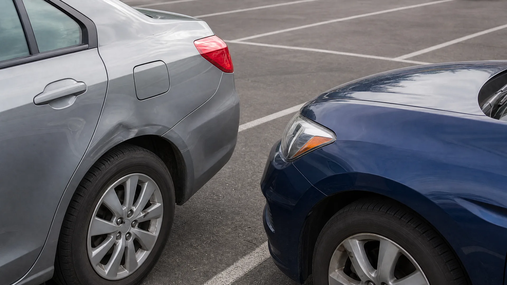 Close-up of a silver car's rear corner and a dark blue car's front corner nearly touching in a paved parking lot with painted white lines.