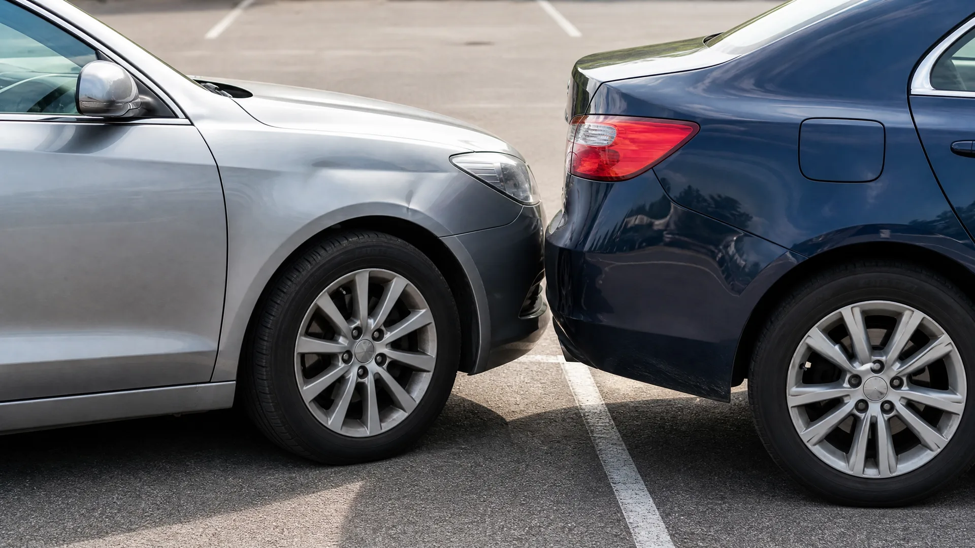 Front end of a silver car touching the rear bumper of a dark blue car in a paved parking lot with painted white lines.