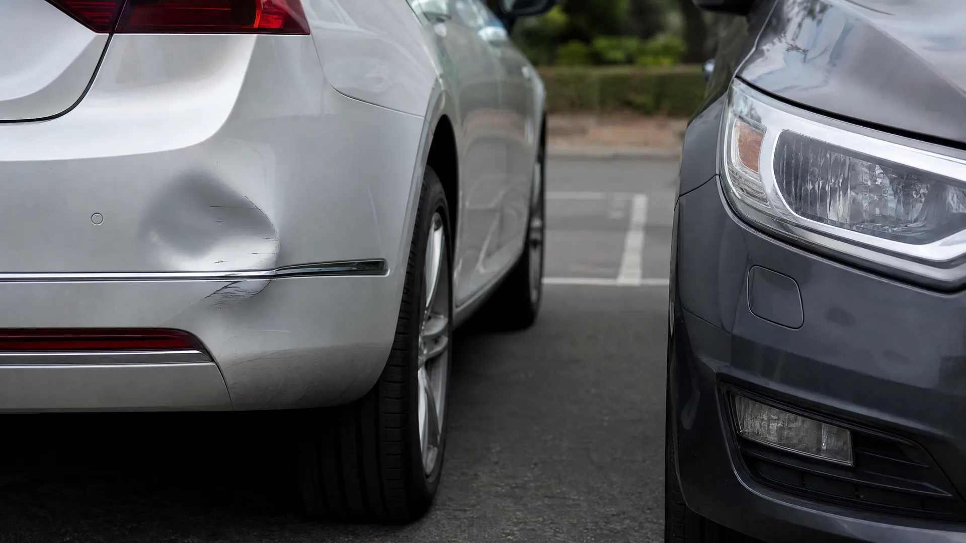 Close-up of a silver car's rear quarter panel with a dent and scrape above the bumper trim, parked next to the front end of a dark gray car on asphalt.