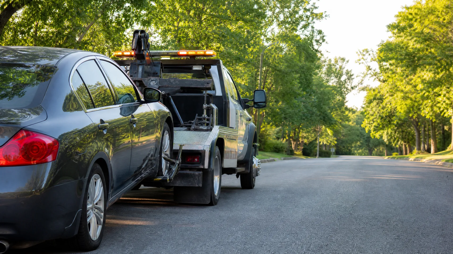 A dark gray sedan loaded onto the flatbed of a tow truck parked on a tree-lined residential street.