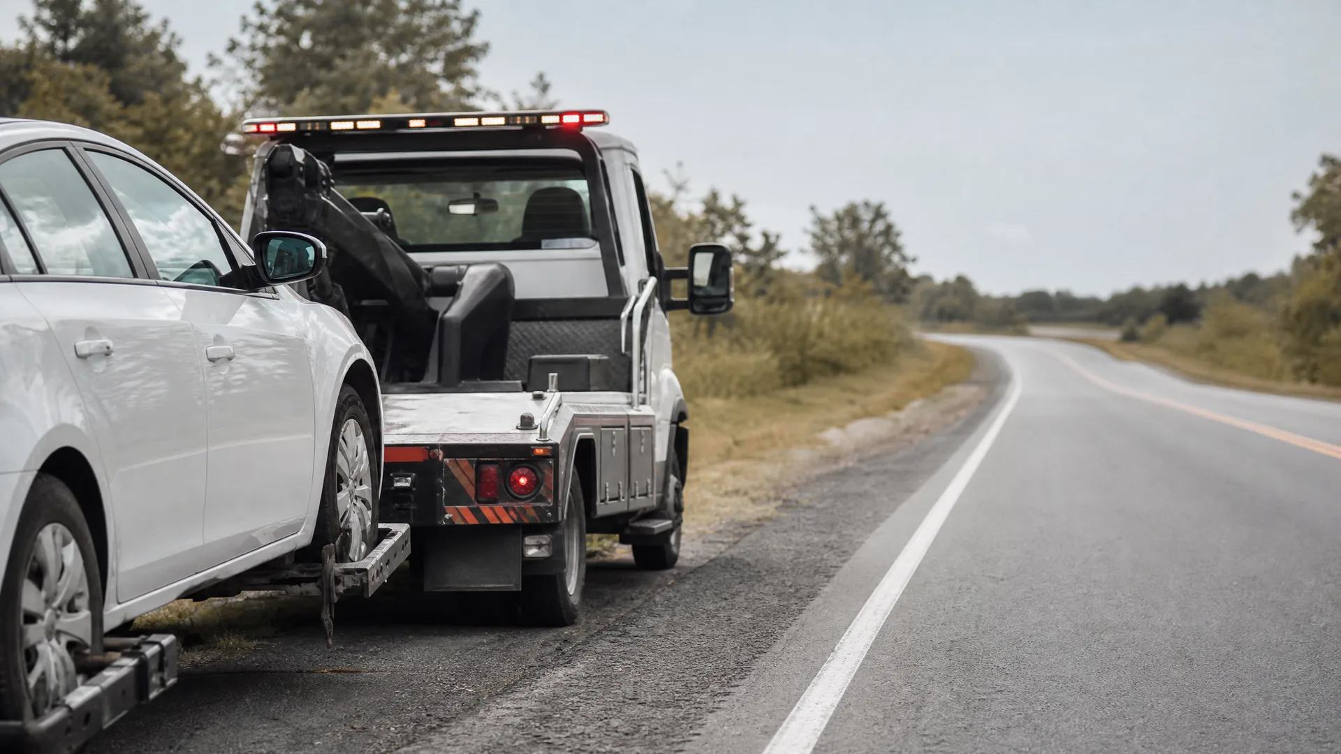A tow truck with a white car hitched to it parked on the shoulder of a two-lane road lined with grass and trees.