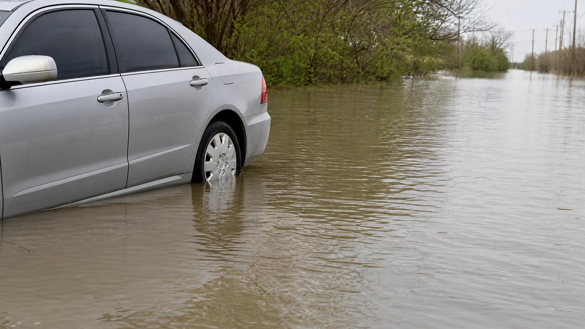 A silver sedan sits in brown floodwater covering a road, with its wheels partly submerged, beside trees and distant utility poles under an overcast sky.