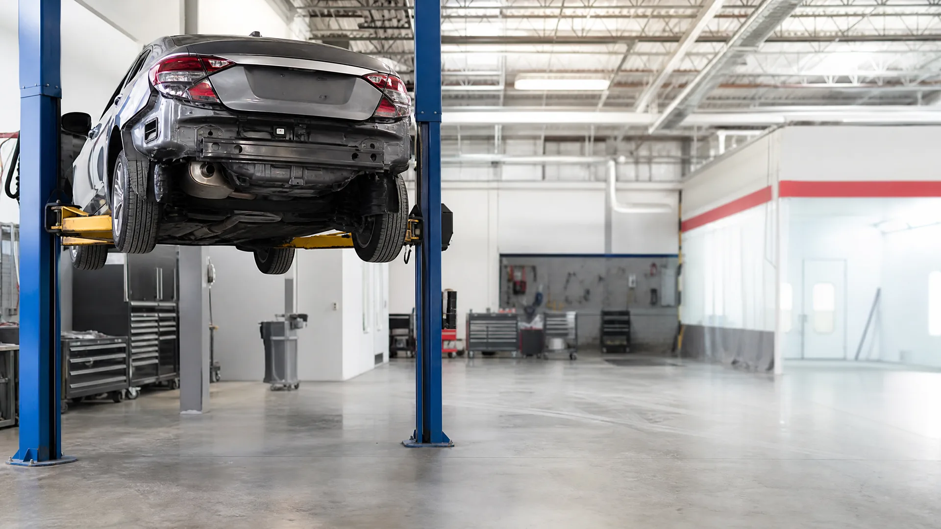 A dark sedan with its rear bumper cover removed is raised on a blue two-post lift inside an auto shop with concrete floors, tool cabinets, and a curtained work bay.