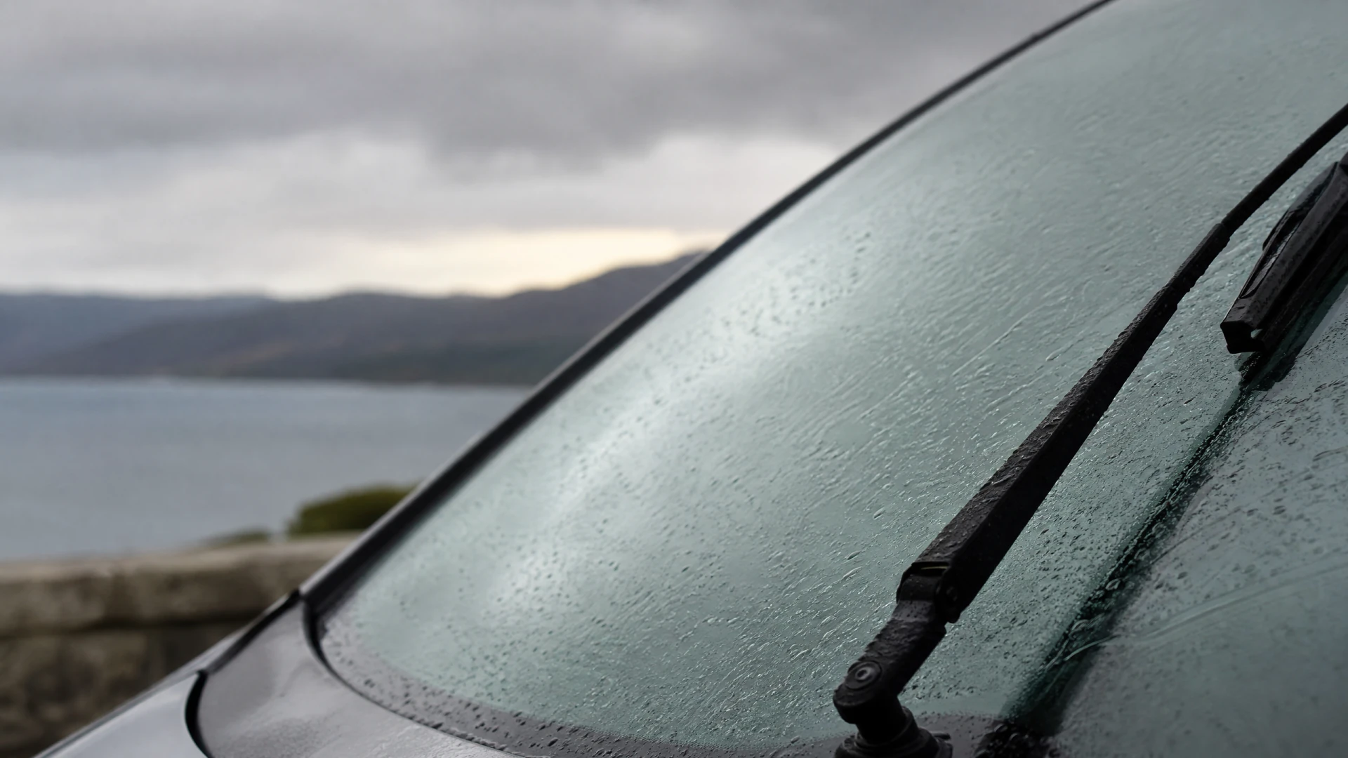 A wet car windshield with a wiper blade, photographed in front of a lake and hills under a cloudy sky.