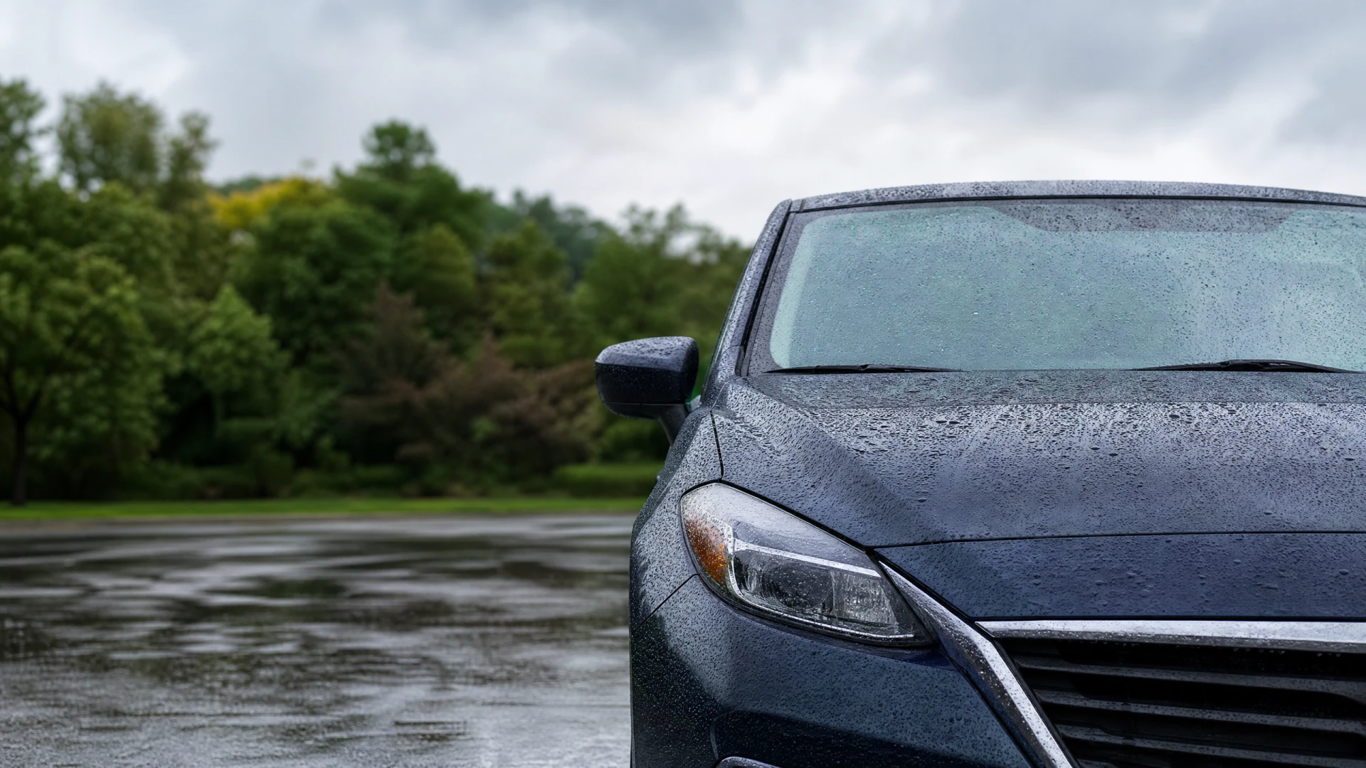 Close-up of the rain-covered hood, windshield, headlight, and side mirror of a dark blue sedan parked on a wet surface.