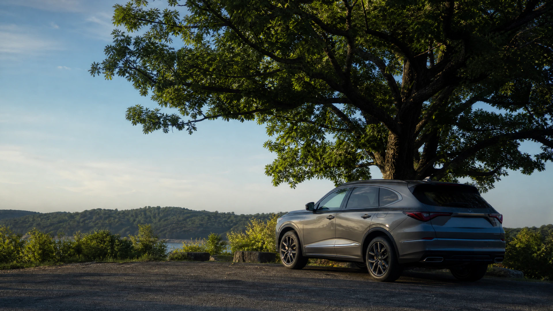A gray SUV parked beneath a large tree on a gravel overlook with a river and forested hills in the background.