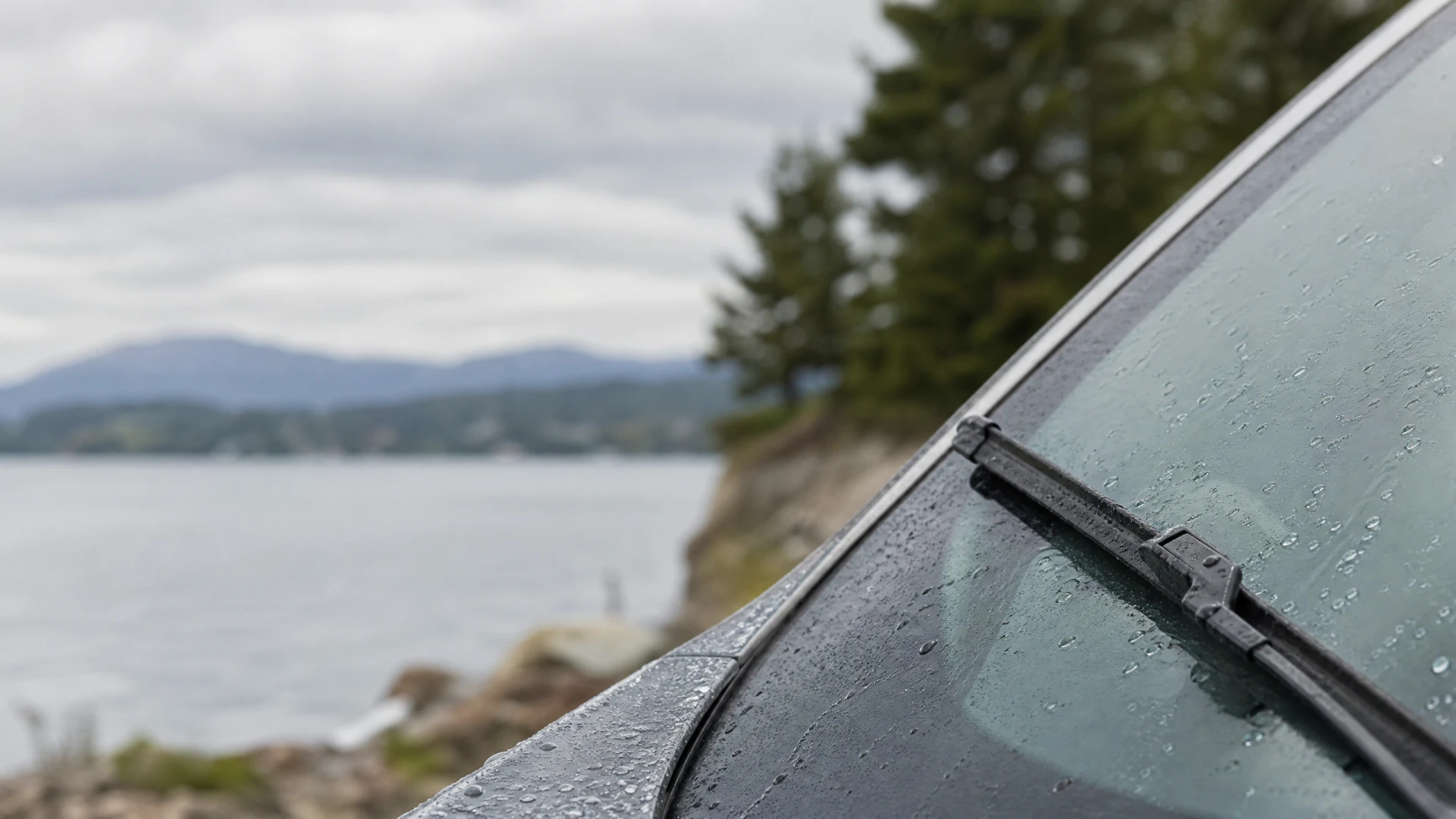 A wet car windshield with a wiper blade, photographed near a lake with mountains in the background.
