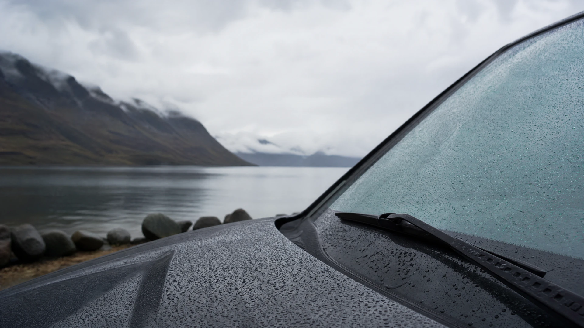 A rain-covered car hood and windshield in the foreground, with a misty mountain lake visible behind it.