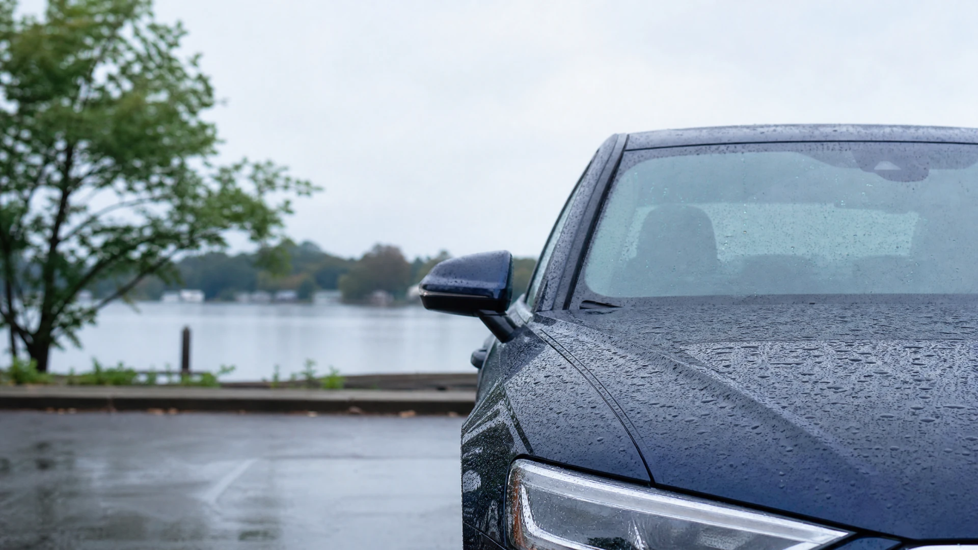 A rain-covered dark blue sedan is parked near a lakeside parking lot on an overcast day.