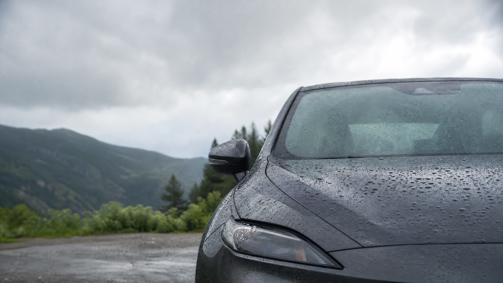 Close-up of a rain-covered dark gray sports car parked in front of a forested mountain range under a cloudy sky.