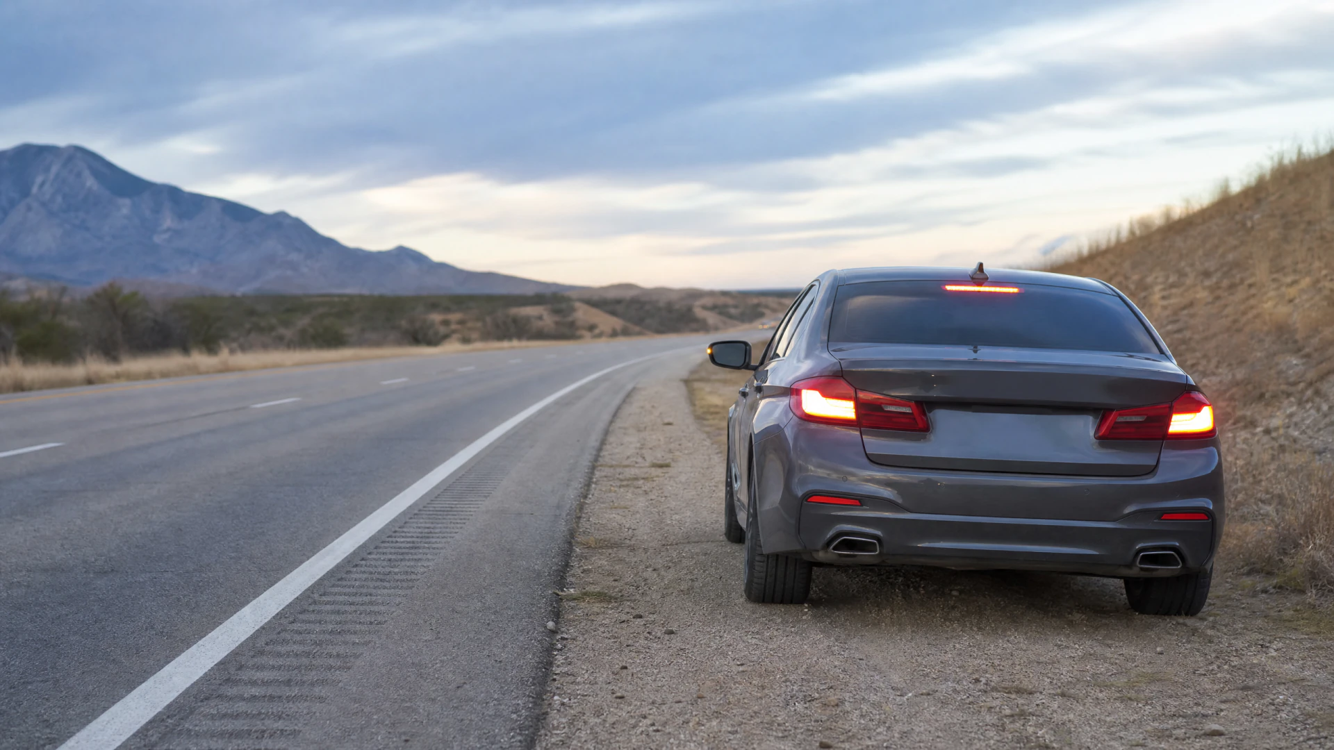 A gray sedan sits on the gravel shoulder of a quiet desert highway with its rear brake lights on, facing away from the camera toward distant mountains.