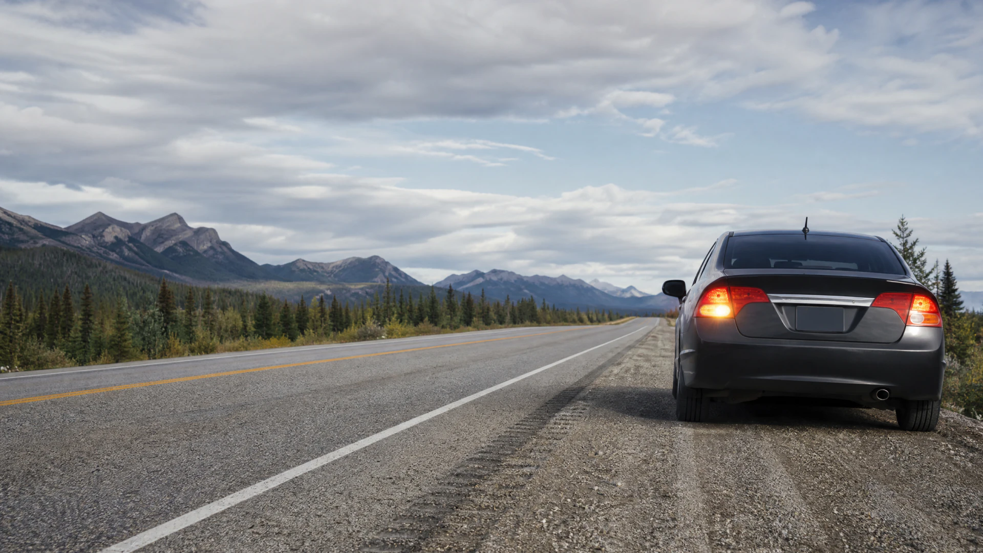 A dark gray sedan sits on the shoulder of a rural two-lane highway surrounded by conifer forests and distant mountains.