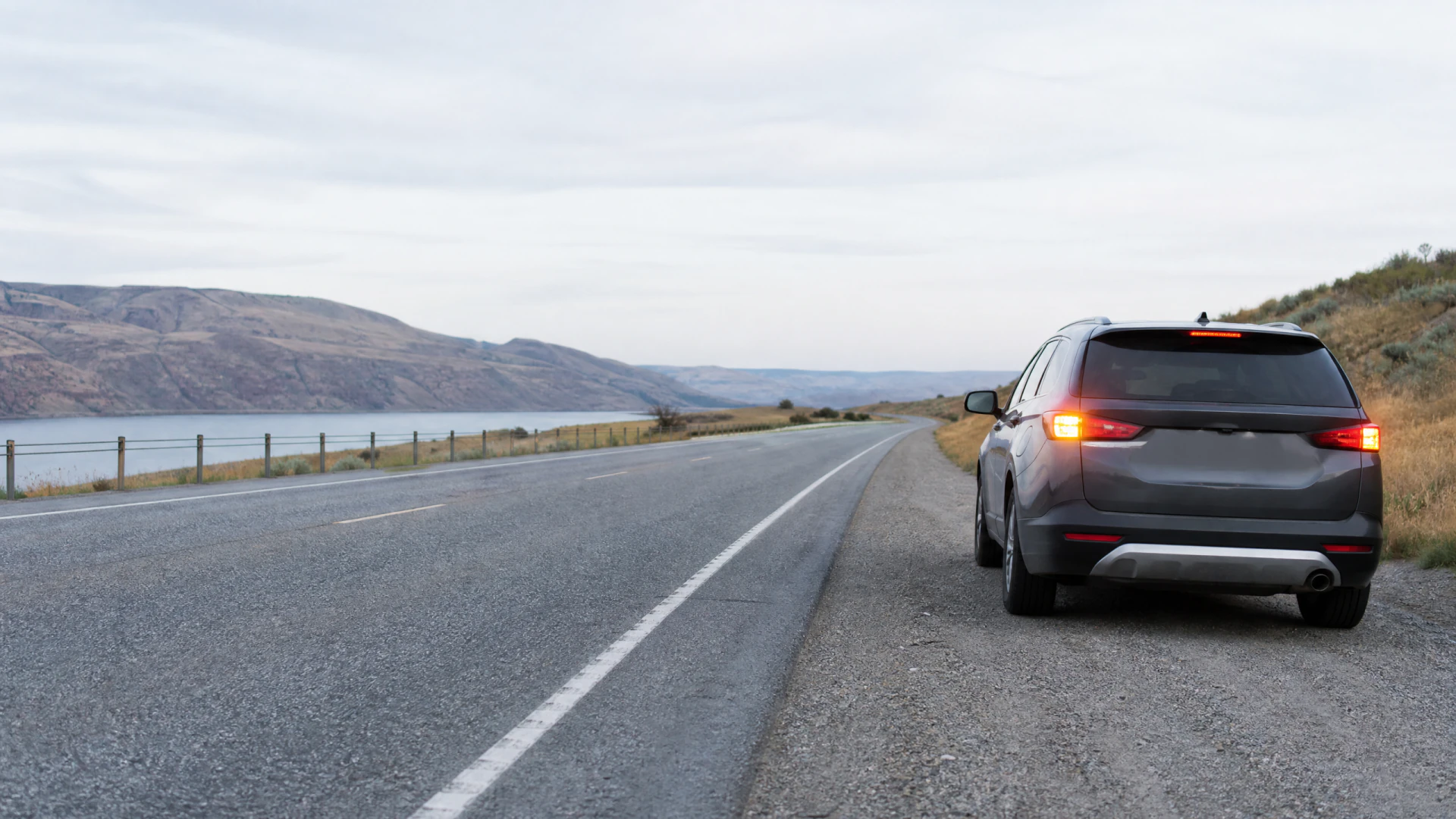 A gray SUV sits on the gravel shoulder of a two-lane highway beside a lake, viewed from behind with its taillights on.