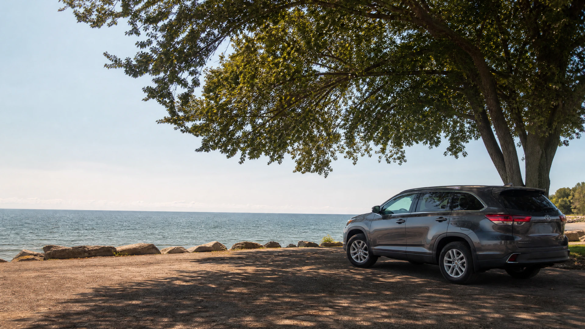 A gray SUV parked in the shade of a large tree beside a calm lake with rocks along the shoreline.
