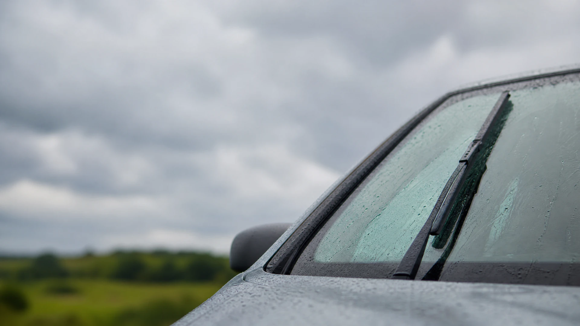 A rain-covered car windshield with a wiper blade, photographed from outside the vehicle under a cloudy sky.