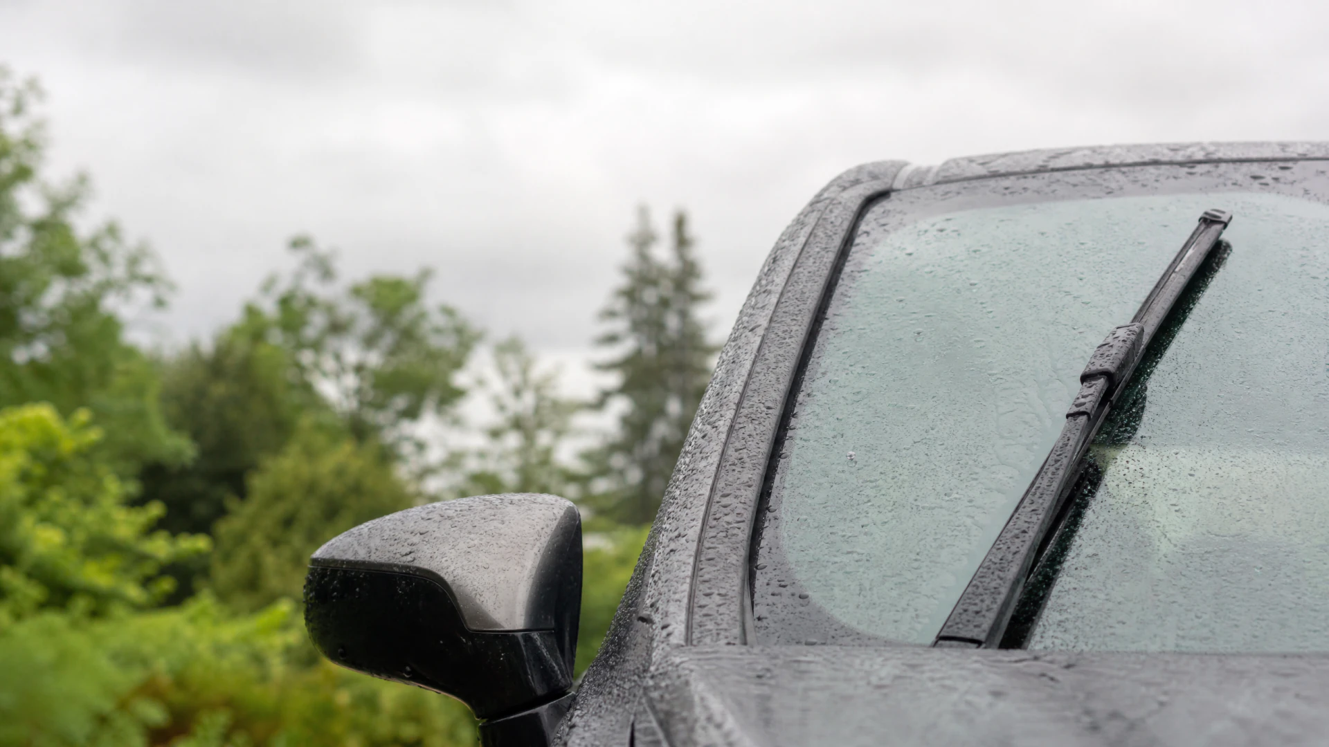 Close-up of a wet dark car windshield with a raised wiper blade, photographed on a rainy day.