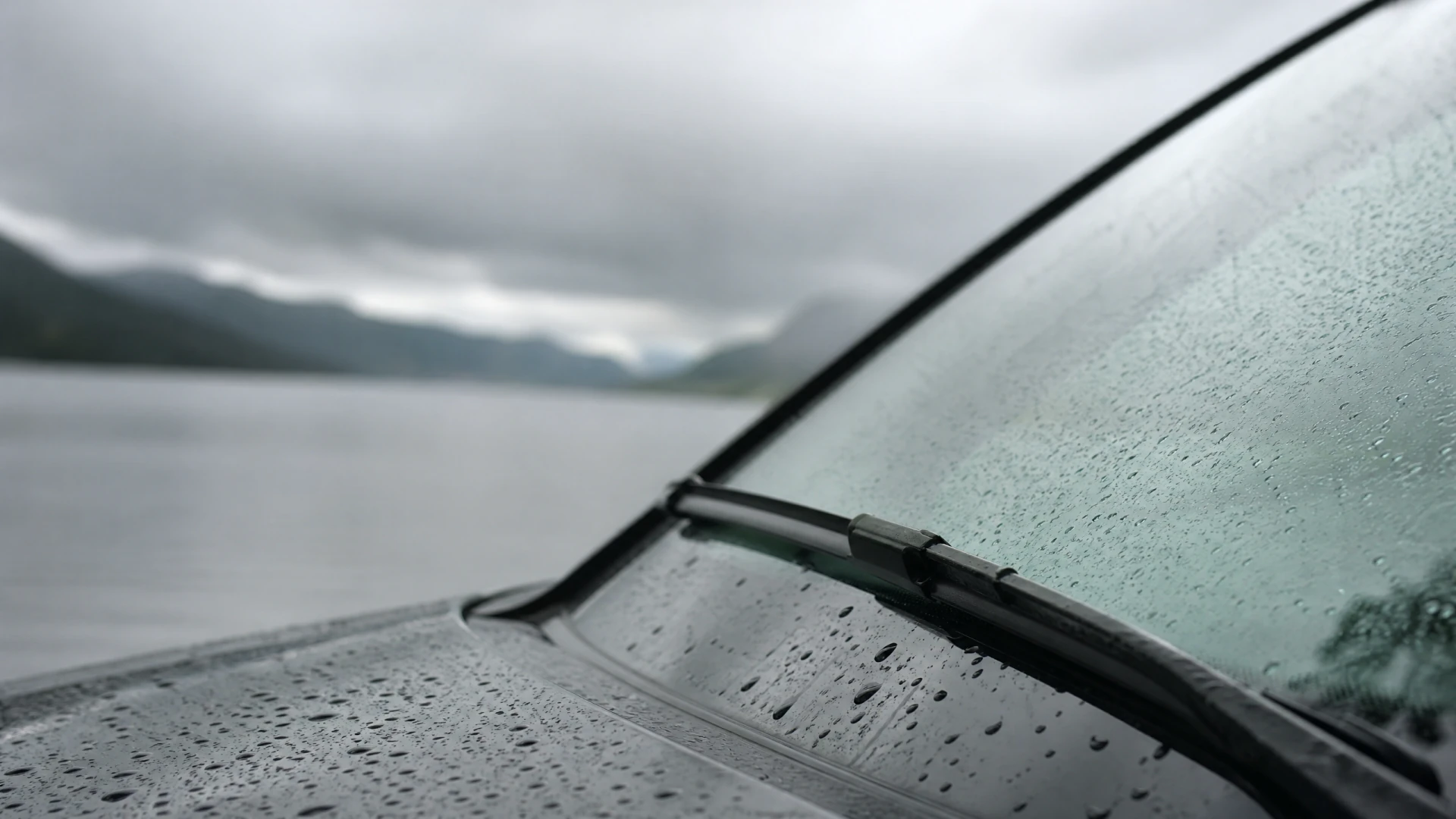 A wet car hood and windshield with a wiper blade, photographed in the rain with a lake and mountains visible in the background.