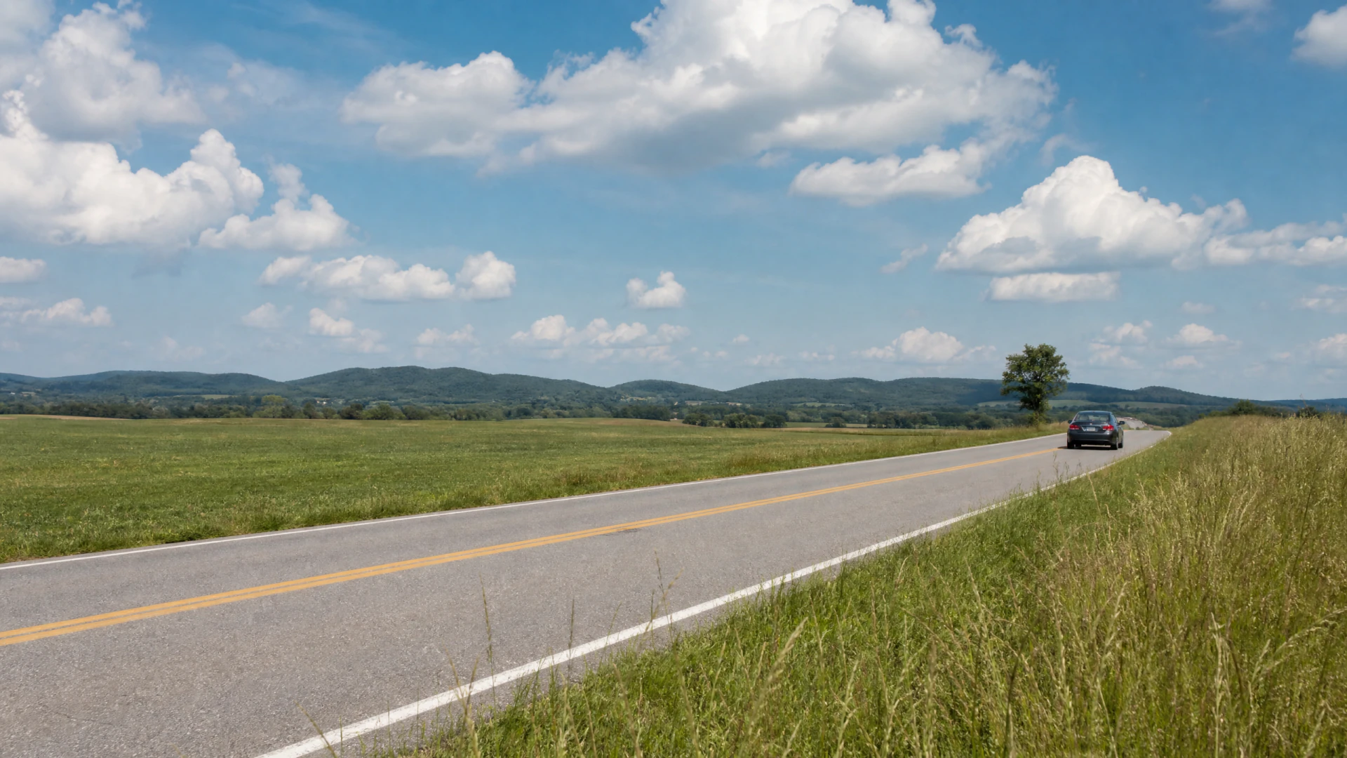A dark sedan drives away from the camera on a two-lane rural road bordered by green fields and low hills.