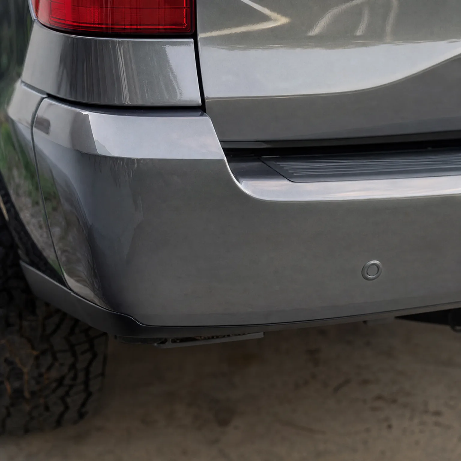 Close-up of the rear corner of a gray SUV showing the lower taillight, bumper with a round parking sensor, and part of the tire above a dirt surface.
