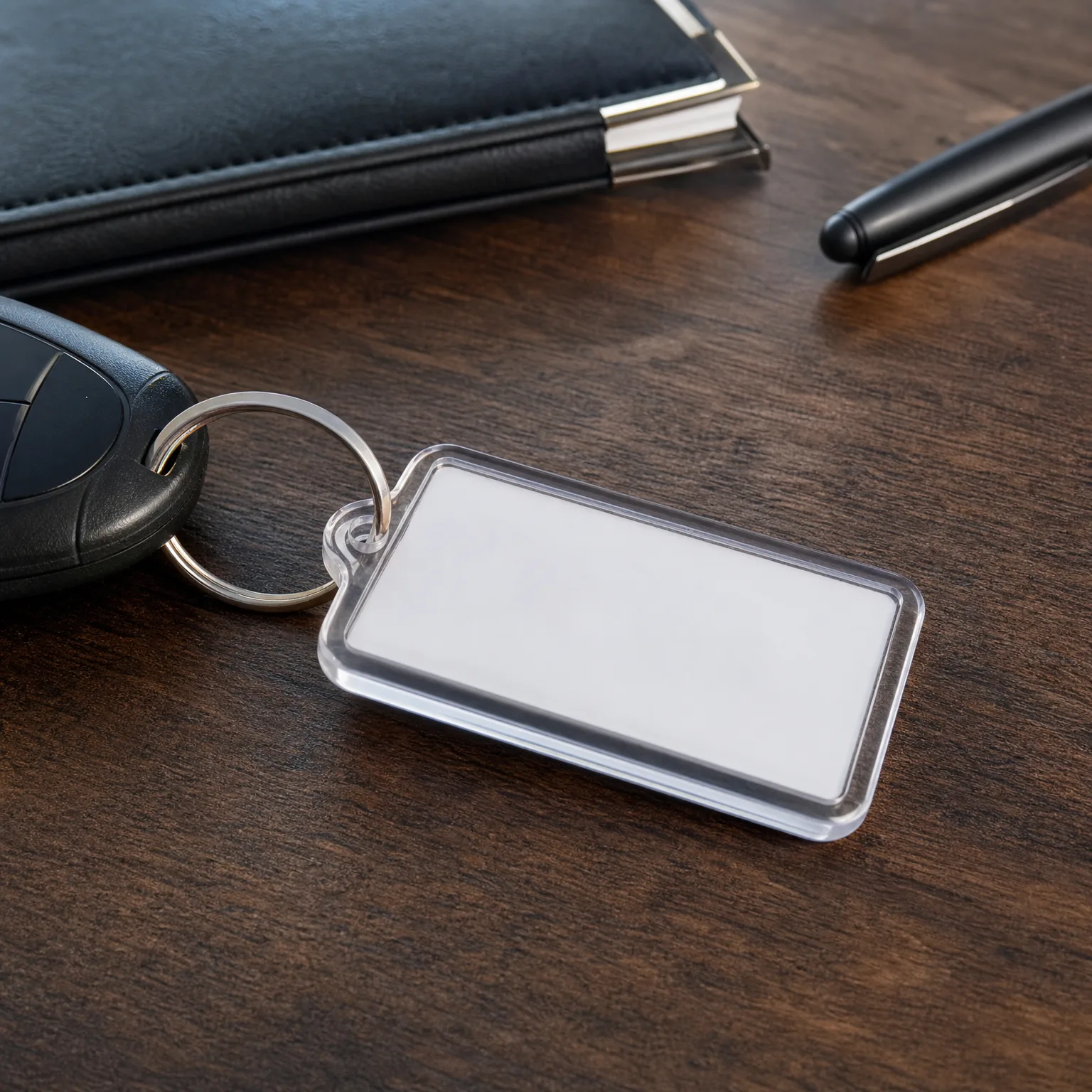 A blank rectangular clear acrylic keychain on a split ring attached to a black car key fob, lying on a dark wood surface beside a black notebook and a black pen.