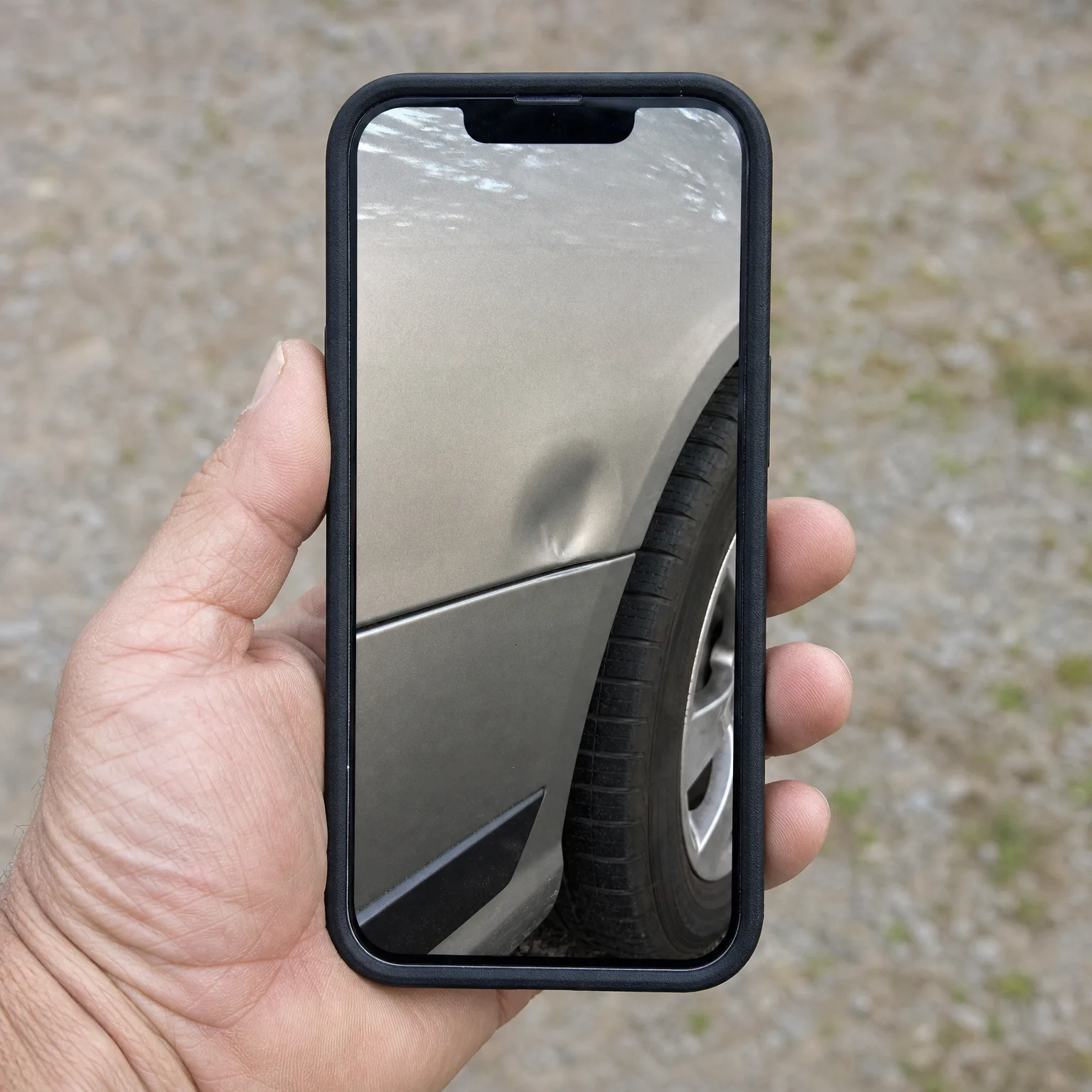 A hand holds a black-cased smartphone displaying a photo of a dent on a silver car's rear fender above the wheel, with blurred gravel ground in the background.
