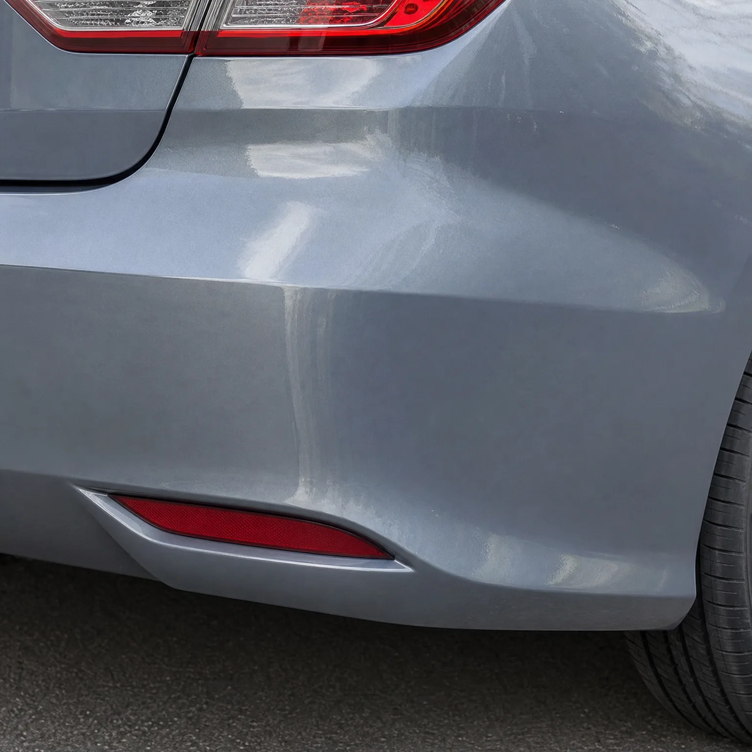 Close-up of the rear corner of a gray car showing a dent and scuffed paint on the bumper near the red reflector, with the taillight and tire edge visible.