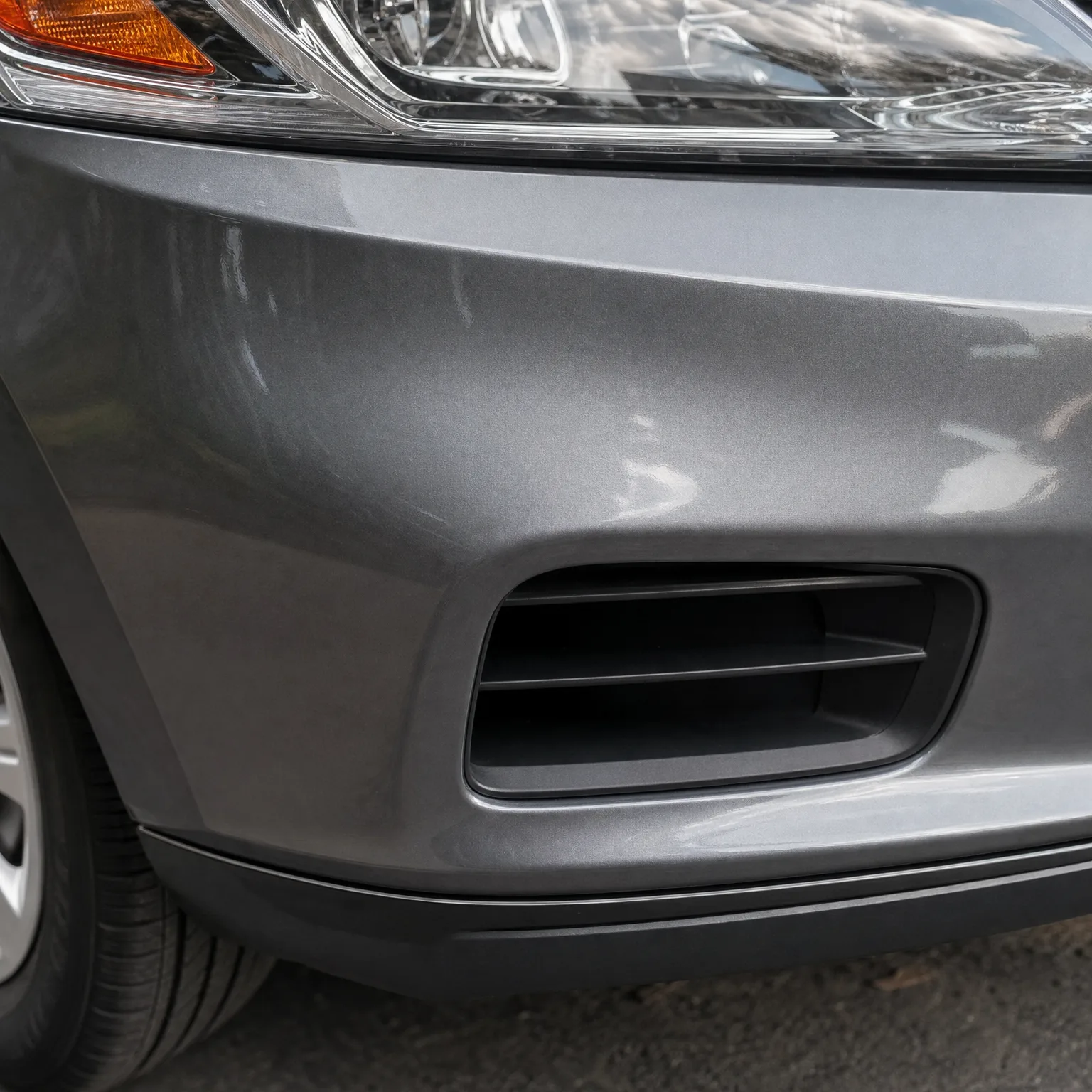 Close-up of the front left corner of a gray car, showing the bumper with a black fog light vent cover, part of the headlight and amber turn signal, and the tire above asphalt pavement.