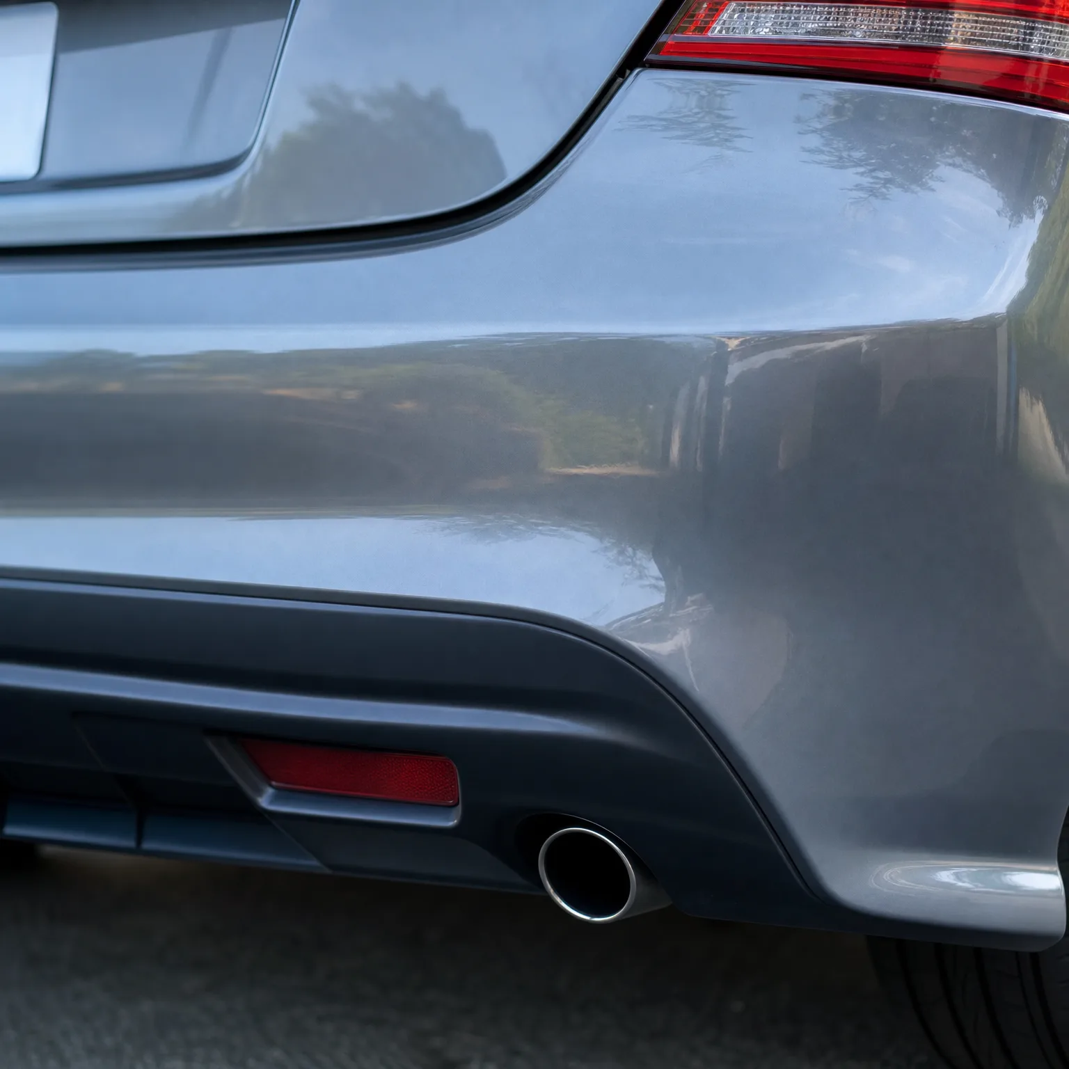 Close-up of the rear bumper corner of a gray car, showing a red reflector, chrome exhaust tip, taillight edge, and trees reflected in the paint.