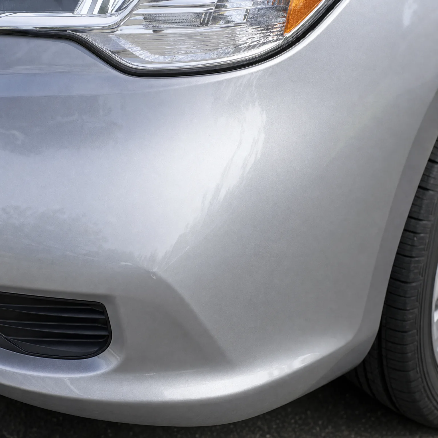 Close-up of the front left corner of a silver car, showing a shallow dent and crease in the bumper below the headlight, with the tire and asphalt at right.