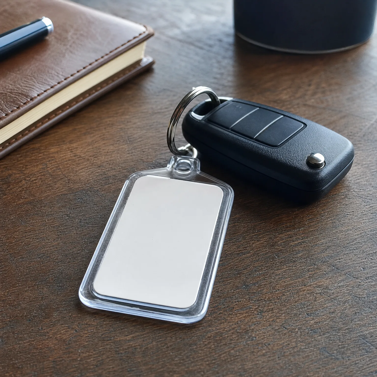 A blank rectangular acrylic keychain attached by a split ring to a black flip-style car key fob, resting on a dark wood surface beside a brown leather notebook and a pen.