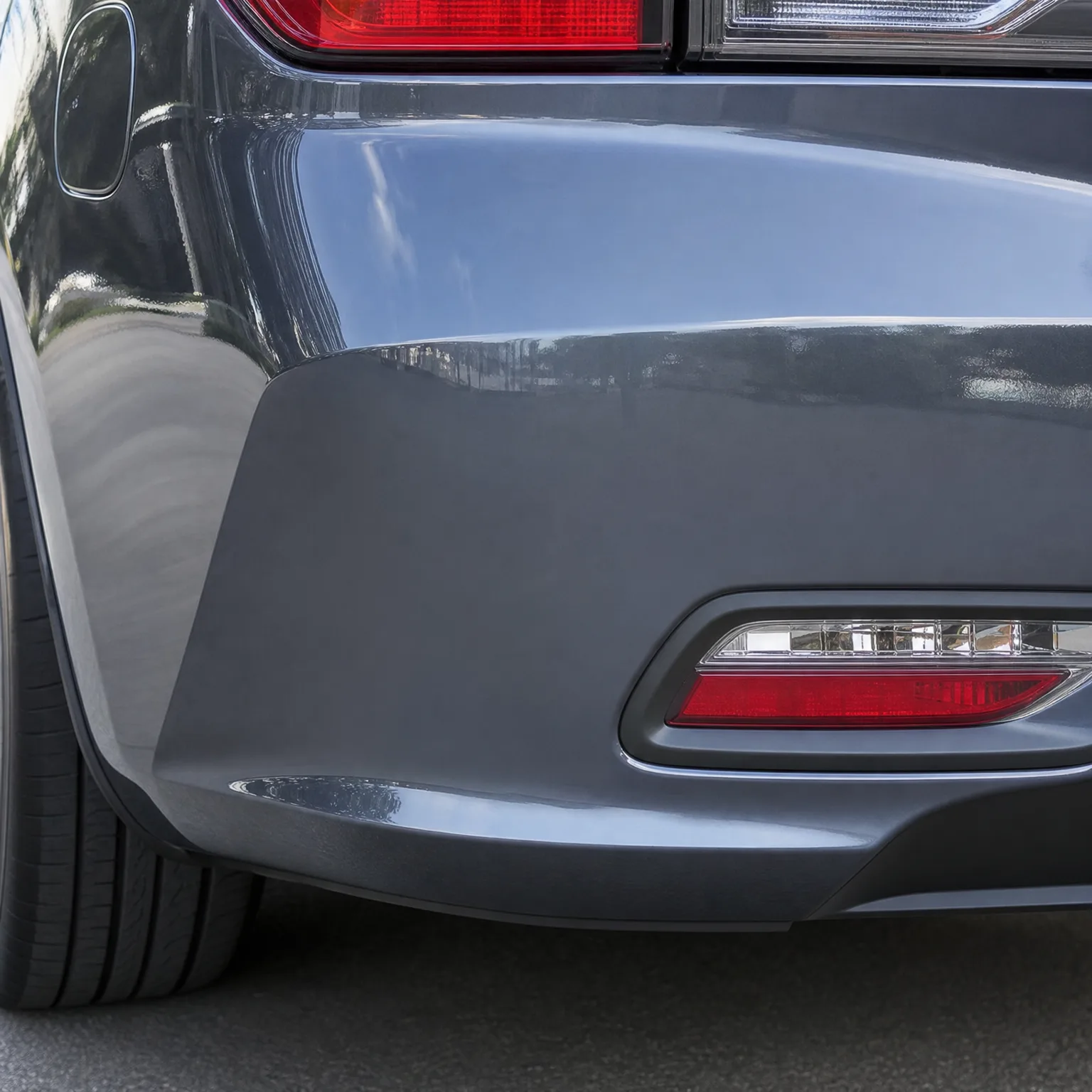 Close-up of the rear corner of a gray car showing the bumper, red reflector with clear lens, part of a taillight, and the rear tire above pavement.